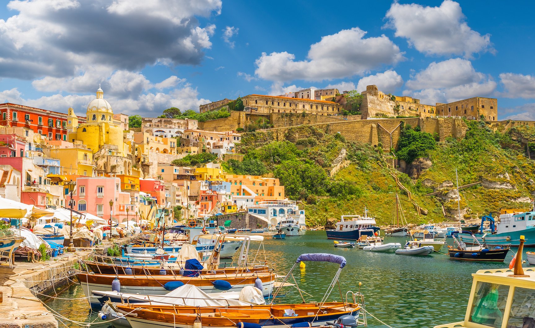 Colorful boats docked in a harbor with a hillside town featuring colorful buildings and a fortress under a partly cloudy sky.