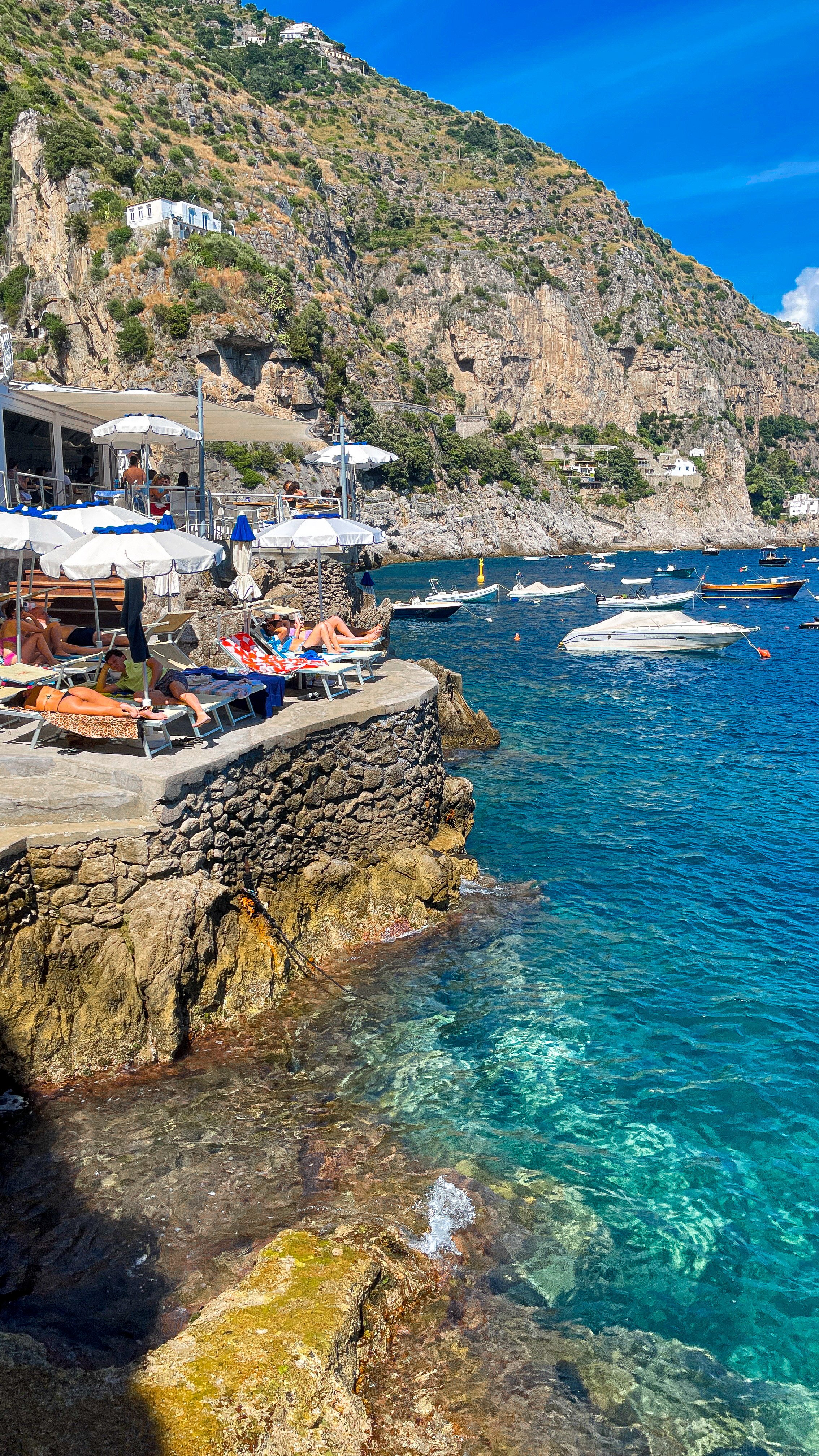 People relaxing on lounge chairs under umbrellas by a rocky waterfront, with boats floating in clear blue water, and a steep hillside with buildings in the background.