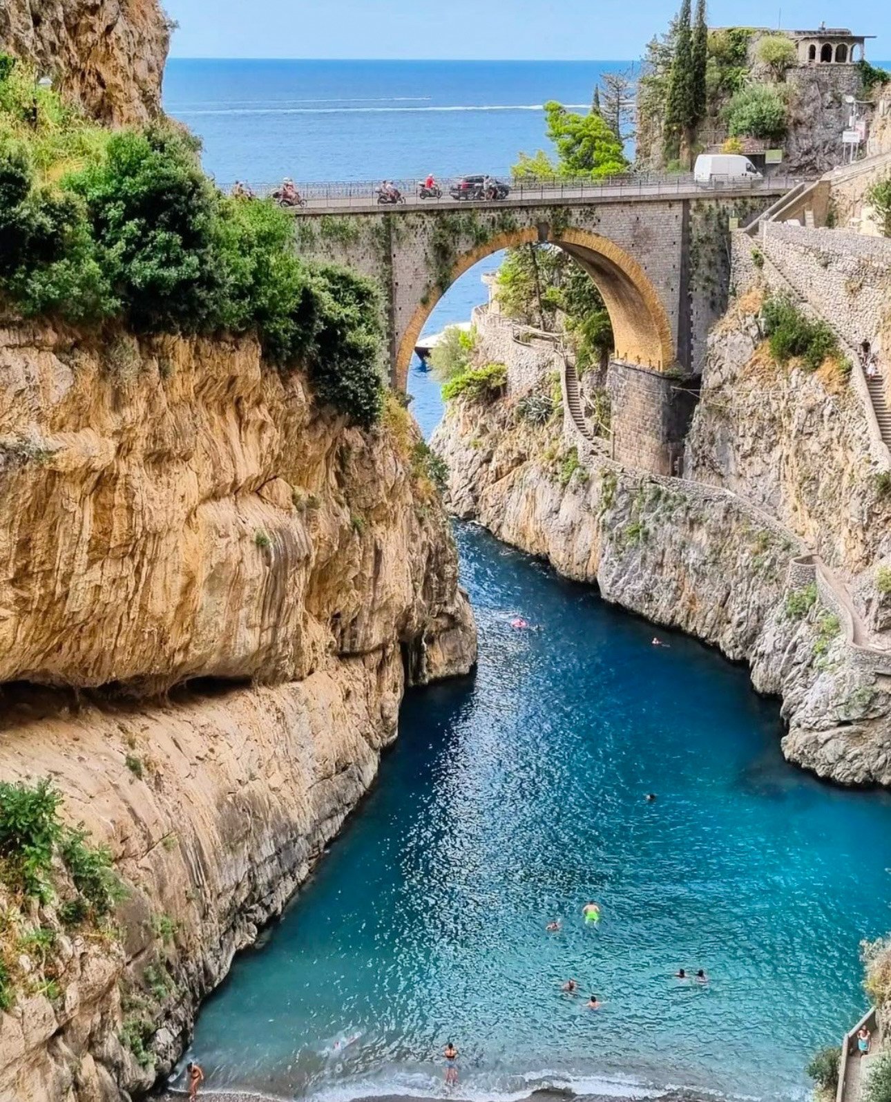 A scenic coastal landscape featuring a stone bridge with cars crossing over a narrow, rocky canyon with a blue waterway. Swimmers are enjoying the water below, and the background shows the sea with a boat passing by.