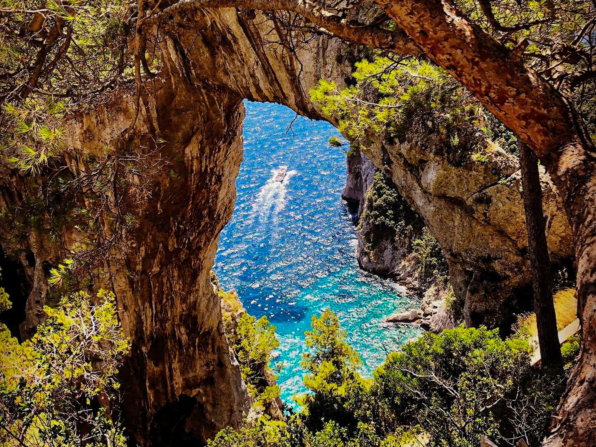 View through a natural rock arch framing a boat on a blue, sparkling ocean with green trees along the rocky shoreline.