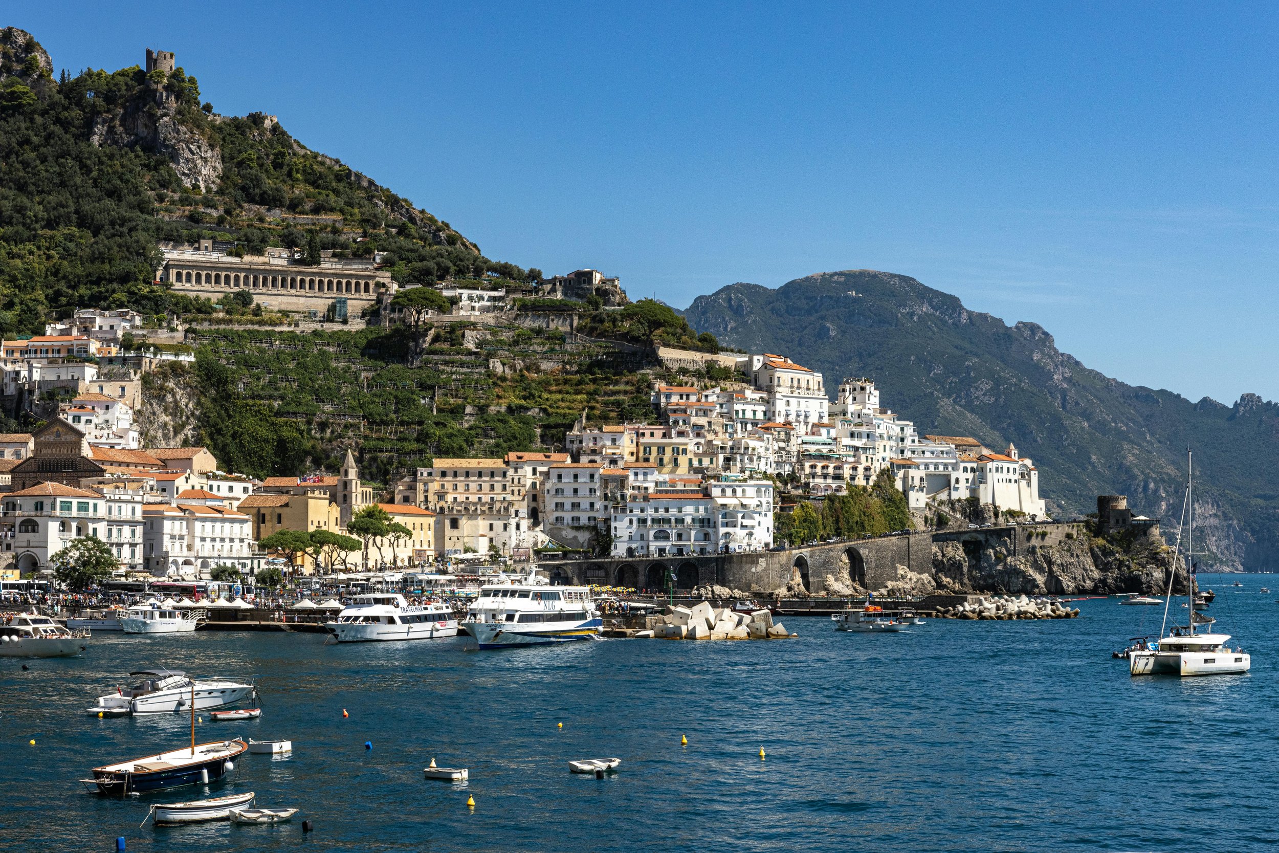 Coastal town with white buildings on cliffs, boats docked in the harbor, mountains in the background, clear blue sky.