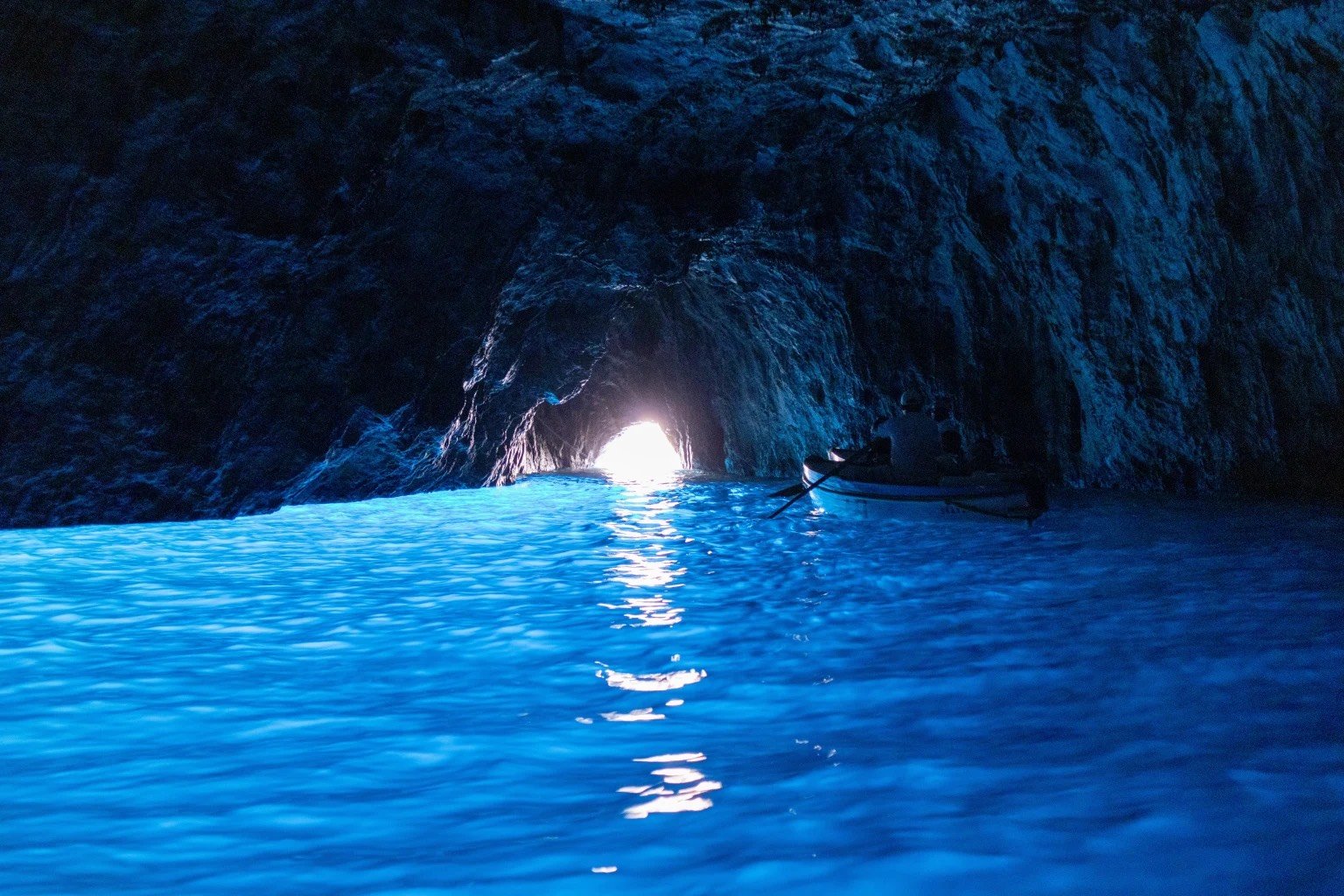 A person kayaking inside a dark ocean cave with light shining from the cave's opening, illuminating the water in blue.