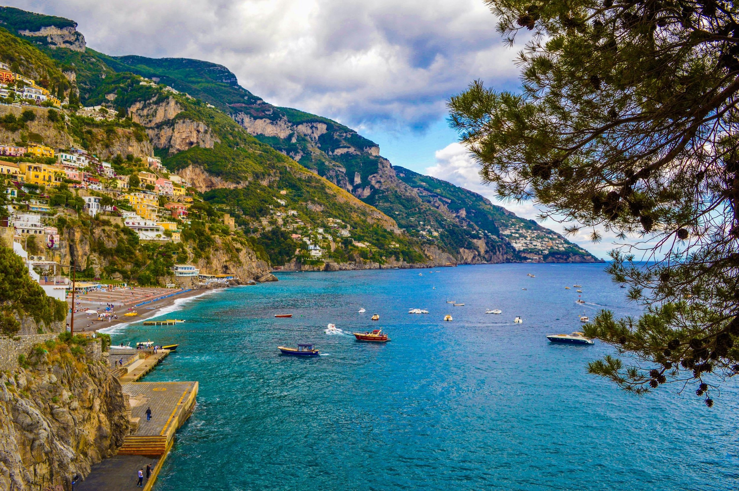 Scenic view of colorful houses along a steep hillside overlooking the Mediterranean Sea with boats anchored near the coast, and a partly cloudy sky.