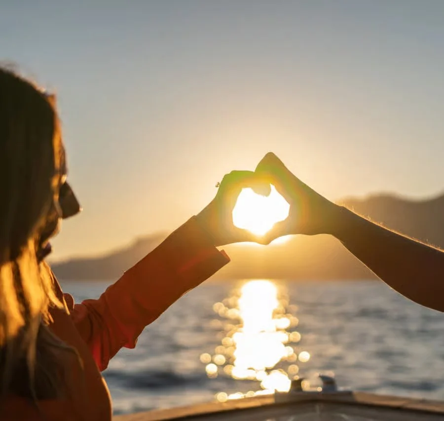 Due persone formano un cuore con le mani davanti a un tramonto sul mare.