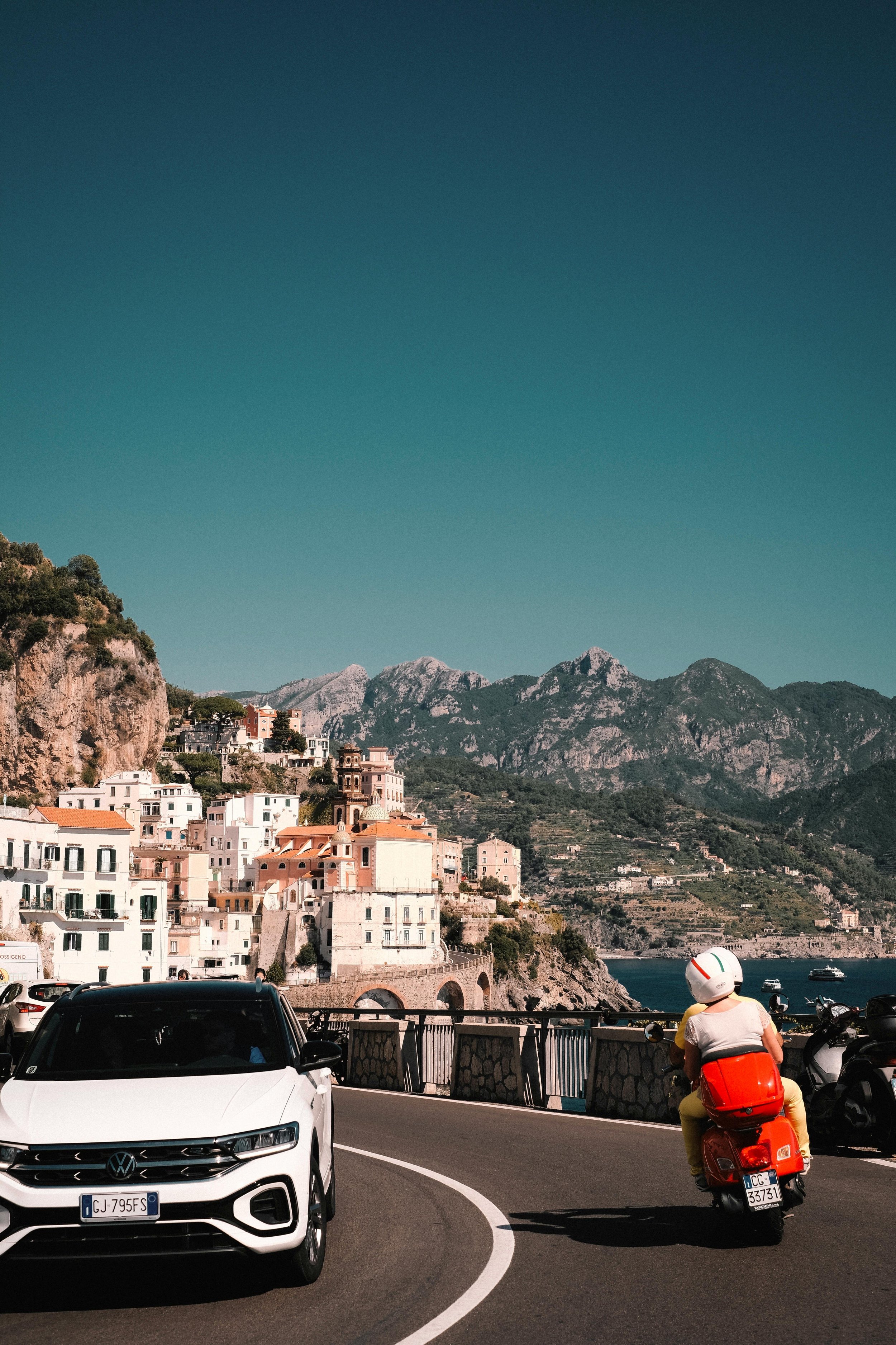 A scenic coastal road with a white Volkswagen car and a person riding a red scooter, overlooking colorful houses on a hillside and mountains in the background under a clear blue sky.
