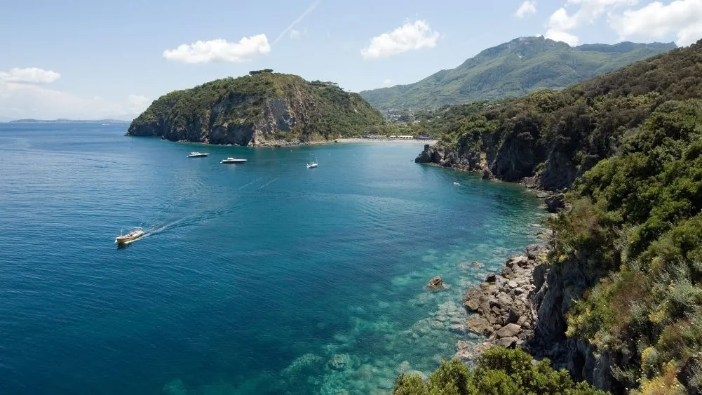 A scenic view of a coastal bay with clear blue water, surrounded by lush green hills and mountains in the background, with several boats sailing on the water.