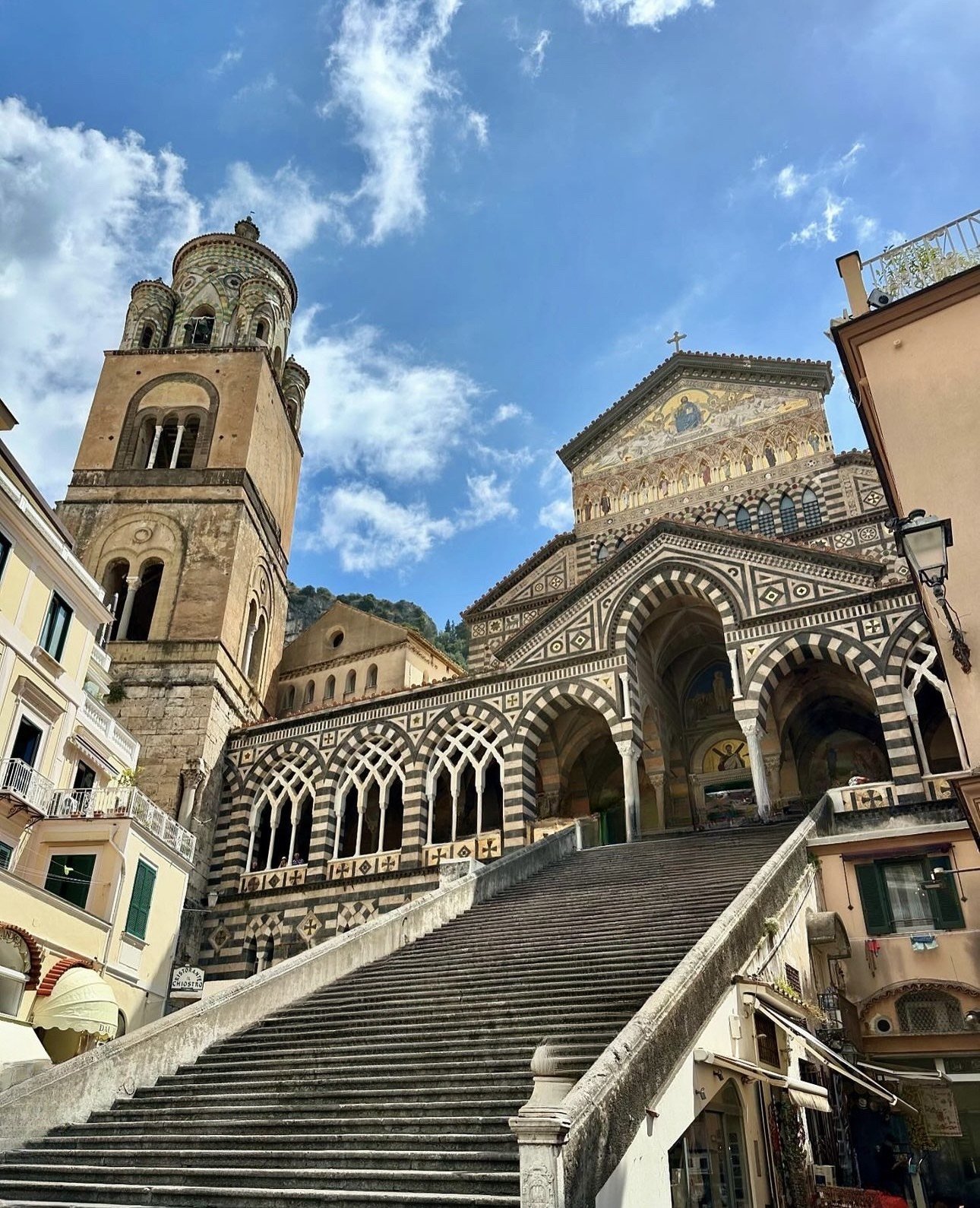 Exterior view of a cathedral with ornate black and white striped facade, a tall bell tower, and a staircase leading up to it, under a partly cloudy blue sky.