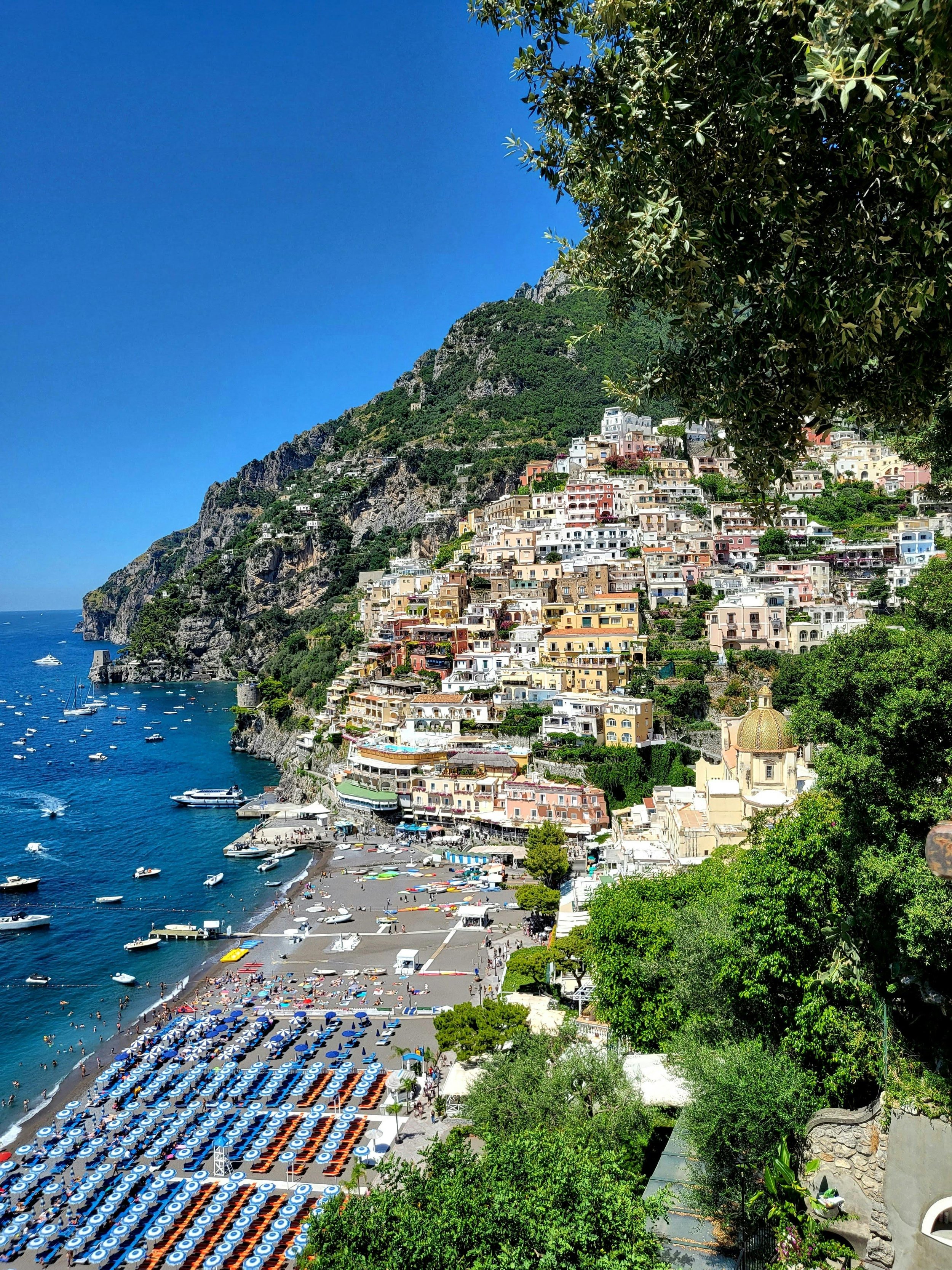 Colorful buildings on a cliffside overlooking a beach with boats in the water, under a clear blue sky, with green trees in the foreground.