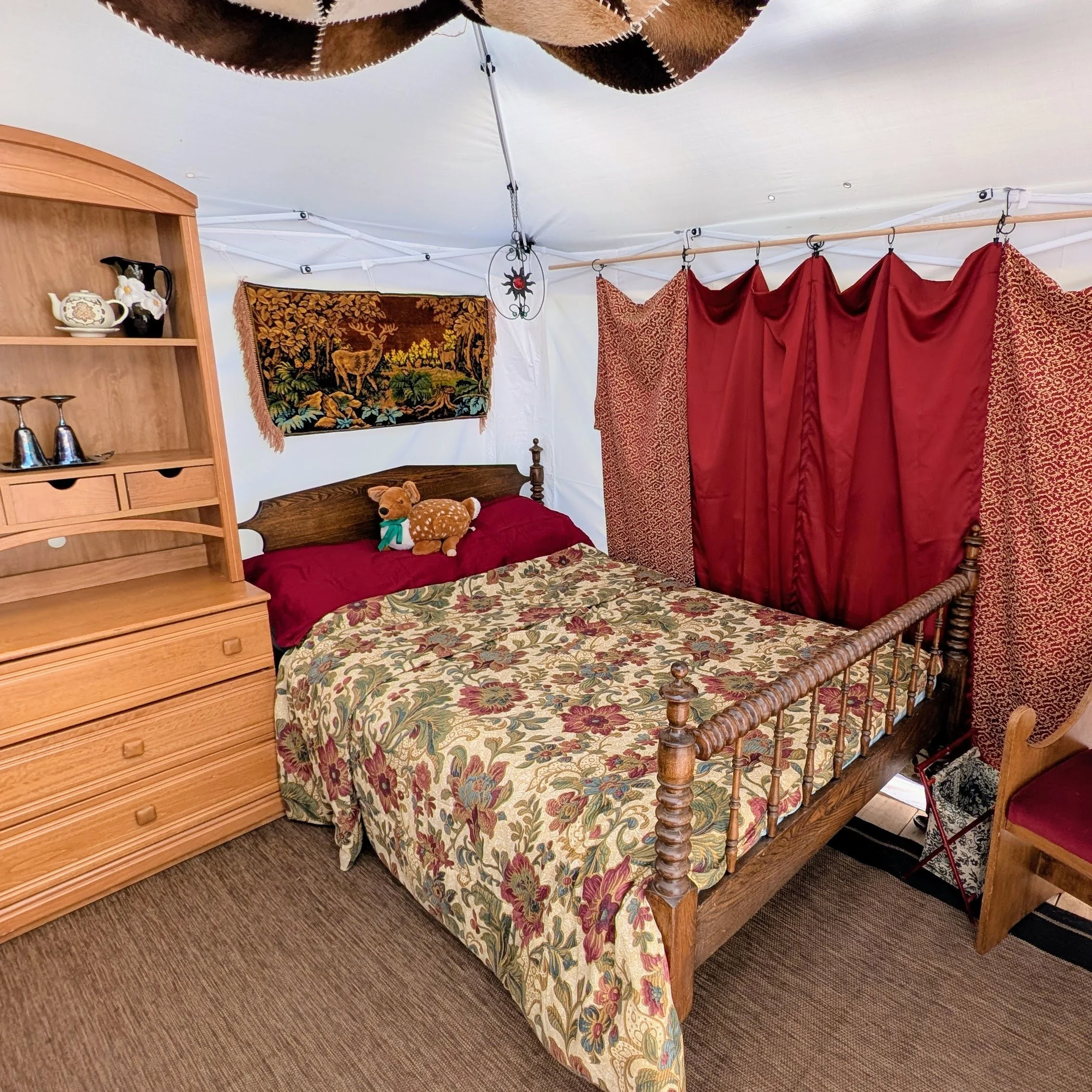 The interior of The Pavilion is outfitted with rich red and gold hued linens and curtains. A deer hide decorative panel hangs above. There is a woven crimson deer tapestry on the wall. The furnishings within the tent are of various shades of wood. Th