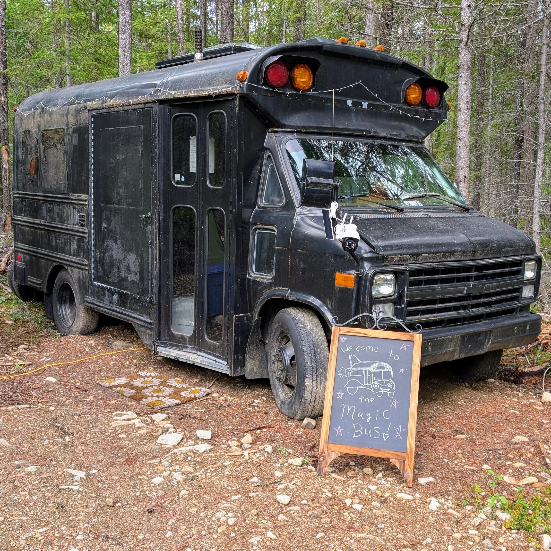 A mini 1989 mini school bus, painted black with twinkle lights hung on it. A small chalkboard in front reads "Welcome to the Magic Bus".