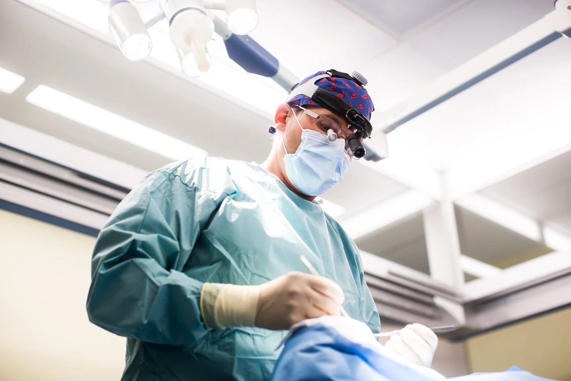 Chirurg in Operationssaal trägt Schutzkleidung, Maske und Spezialbrille, arbeitet an einem Patienten, unter OP-Leuchte.