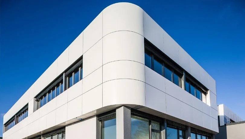 Modernes, weißes Gebäude mit runden Ecken und großen Fenstern unter einem klaren blauen Himmel.