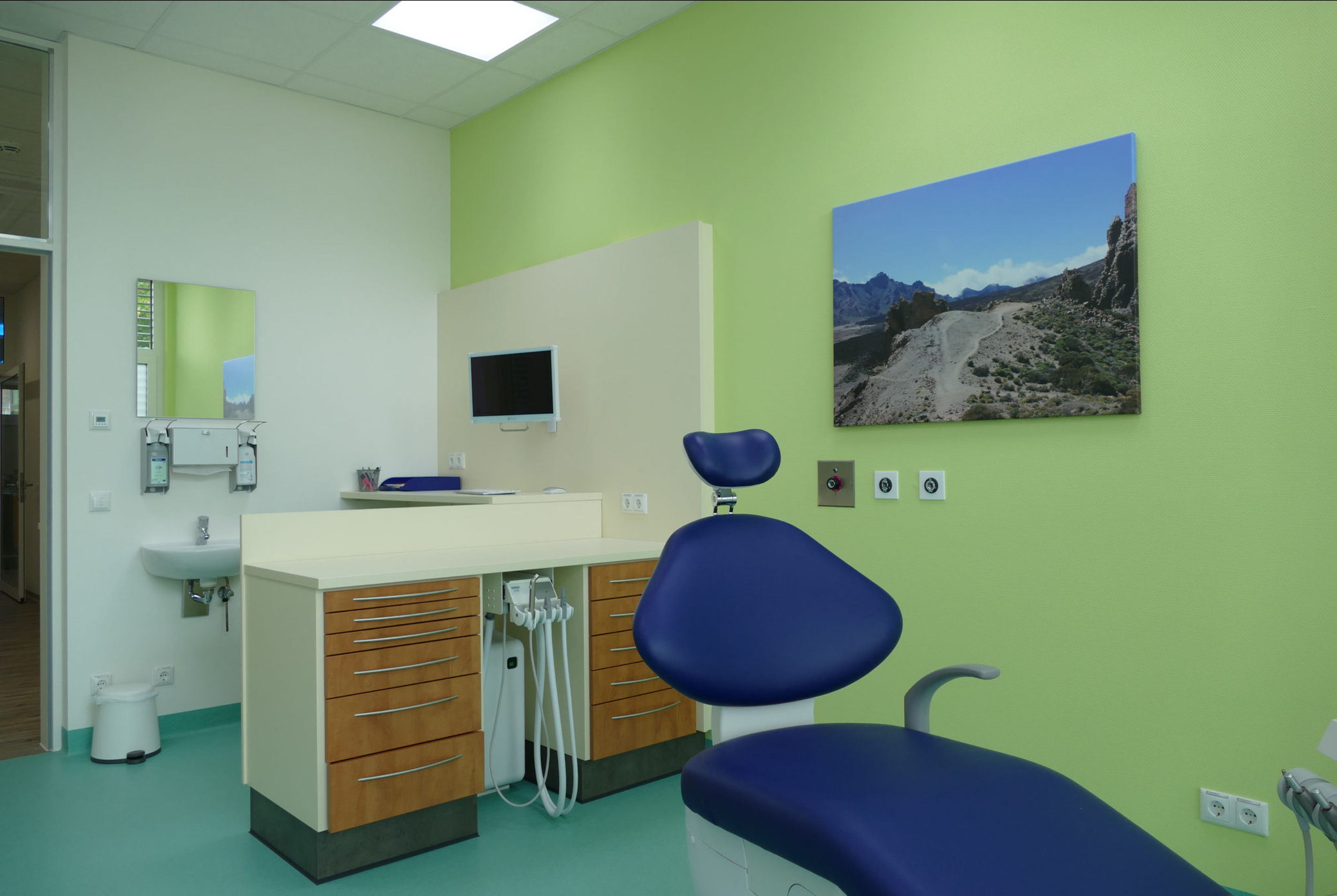 Ein Behandlungszimmer in einer Arztpraxis mit grünen Wänden, einem blauen Behandlungsstuhl, einem weißen Schrank, einem Monitor und einem Wandbild einer Berglandschaft.