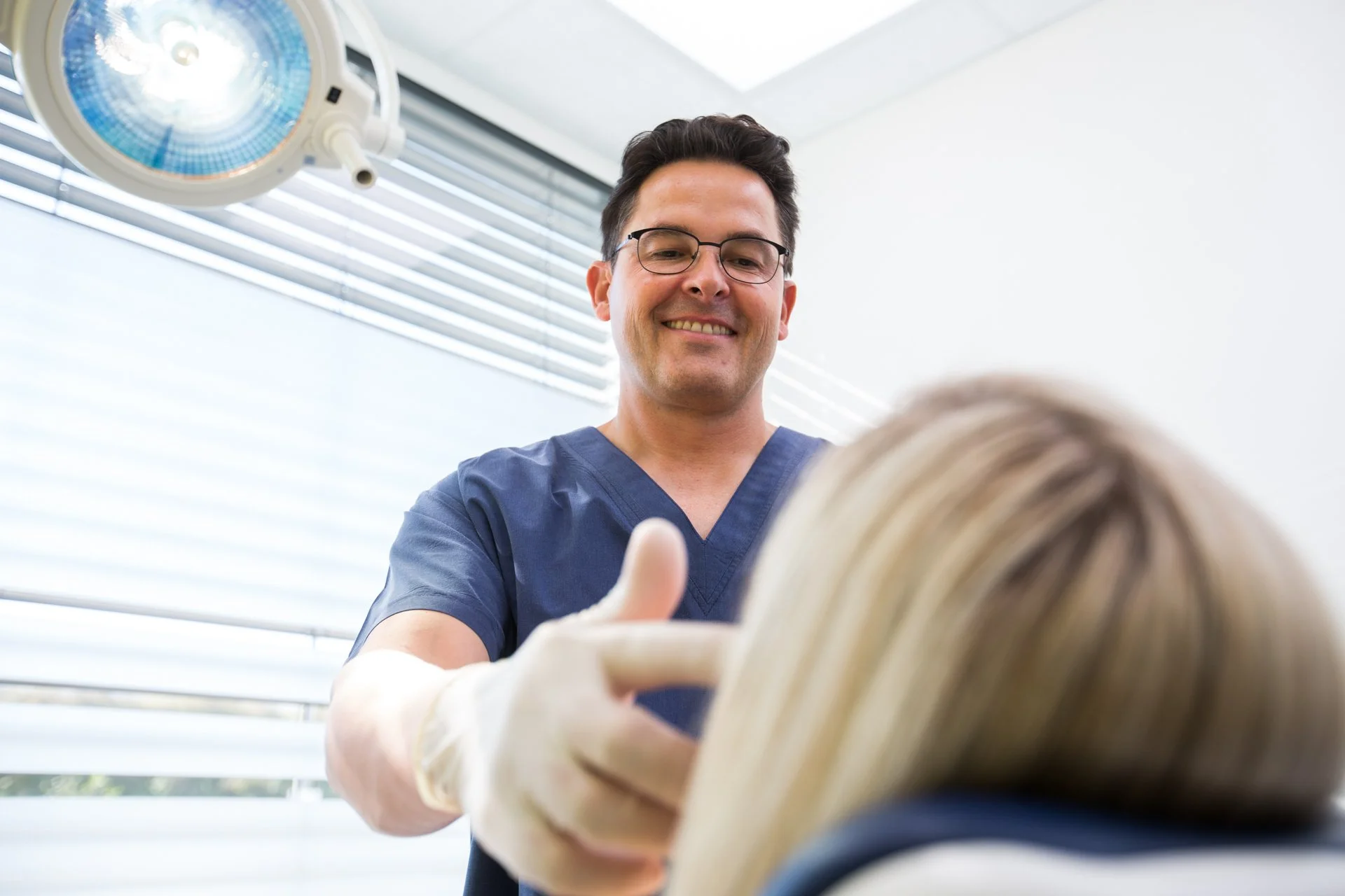 Ein männlicher Arzt in blauer Klinikuniform, der einem Patienten die Hand schüttelt und dabei lächelt, in einem Behandlungsraum mit Operationsleuchte.