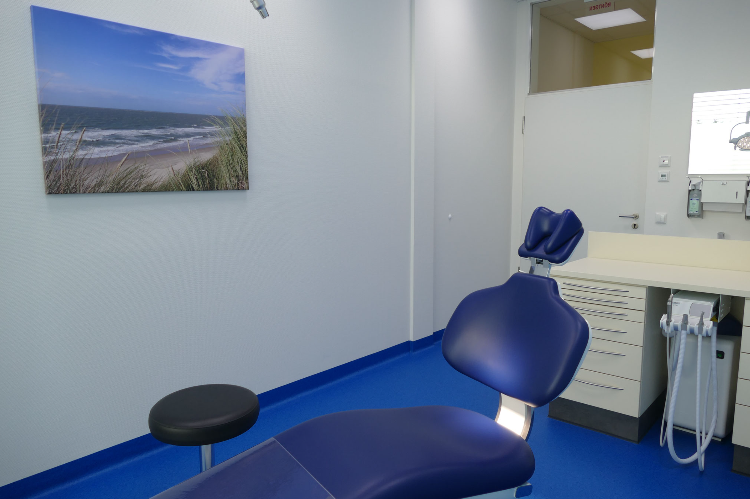 Behandlungszimmer in einer Arztpraxis mit blauer Liege und Stuhl, weißen Schubladen, medizinischem Gerät, Wandbild eines Strandes, weißer Wand und Tür.