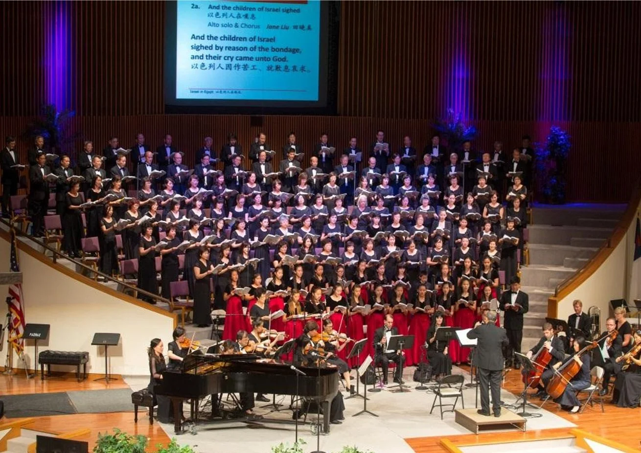 A choir performing on stage with an orchestra in front, with a large screen displaying song lyrics in English and Chinese behind them.
