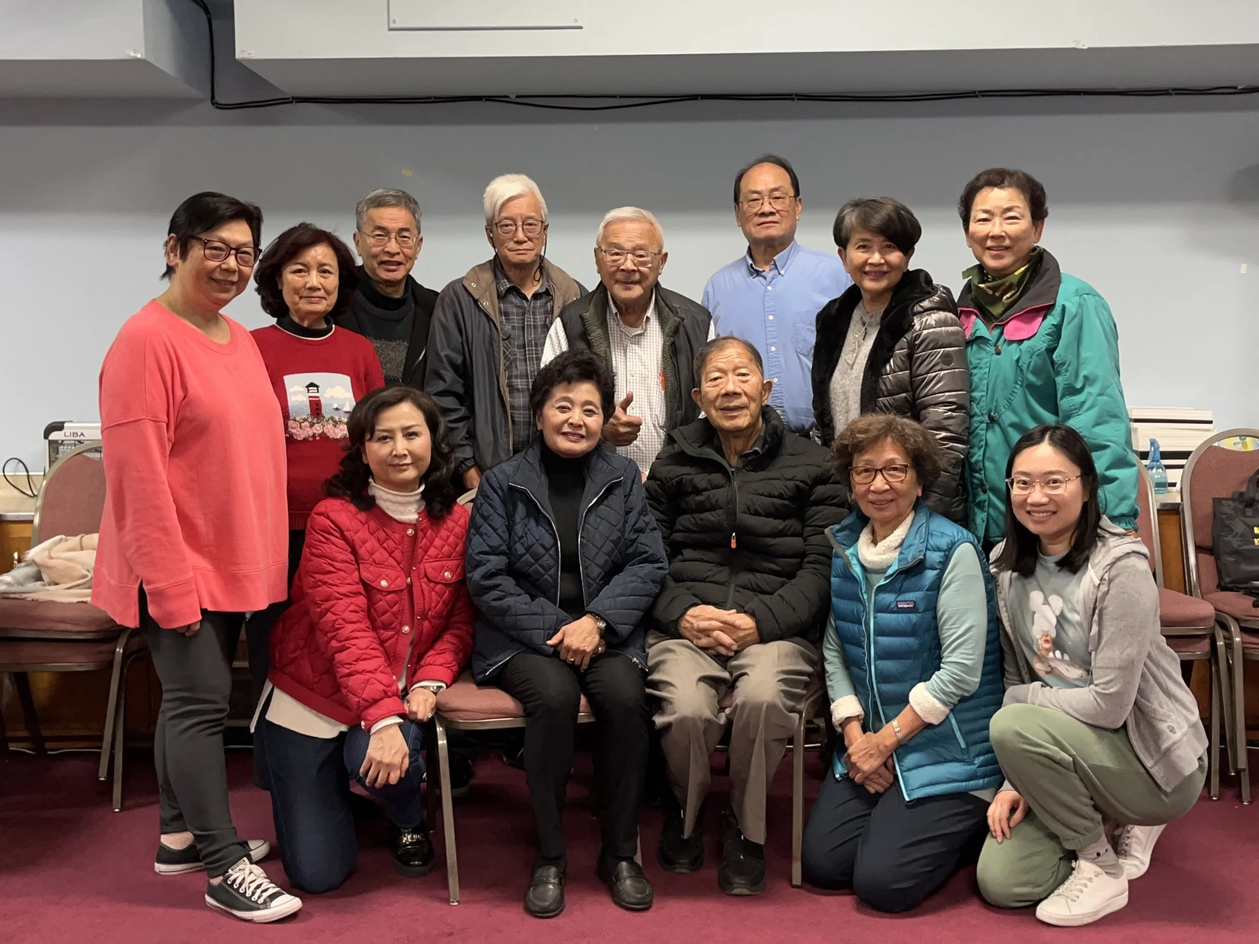 Group of fifteen Asian adults posing indoors for a photo, some seated on chairs and some standing behind, in a room with gray walls and maroon carpet.