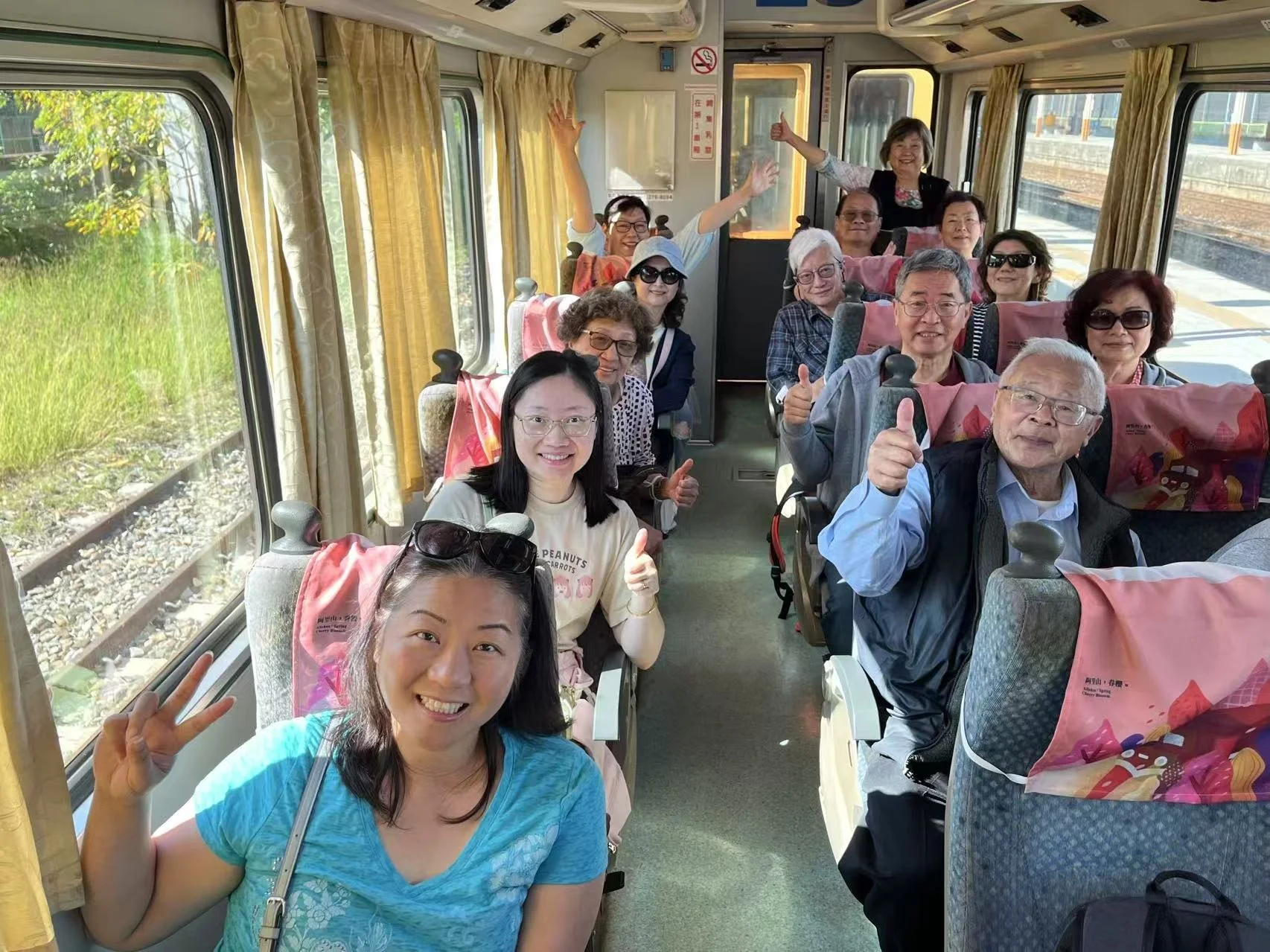 Group of happy elderly and middle-aged people on a train, smiling and giving thumbs-up, with some waving and making peace signs.