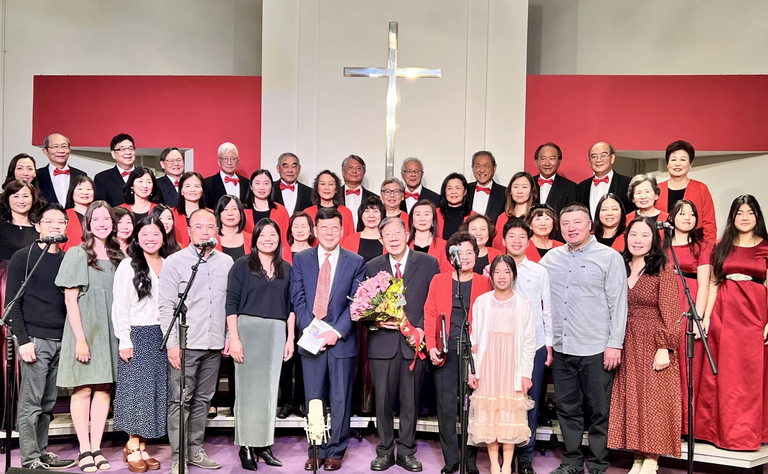 A large choir wearing formal attire, with women in black or red dresses and men in tuxedos with red bow ties, standing on a stage with a cross on the wall behind them, posing for a group photo after a church service or concert.