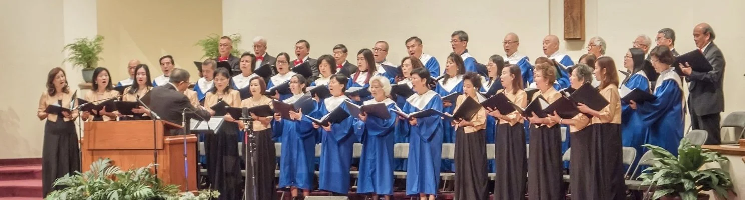 A choir of men and women singing on stage, directed by a conductor, in a church or auditorium setting.