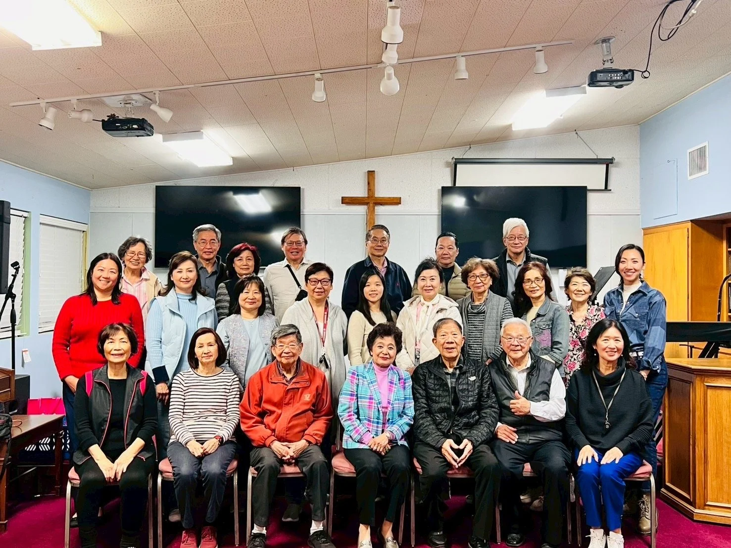 A group of twenty-five diverse people, mostly seniors, posing for a photo inside a church sanctuary with a cross on the wall behind them. They are standing and sitting in front of two large screens and a wooden pulpit.
