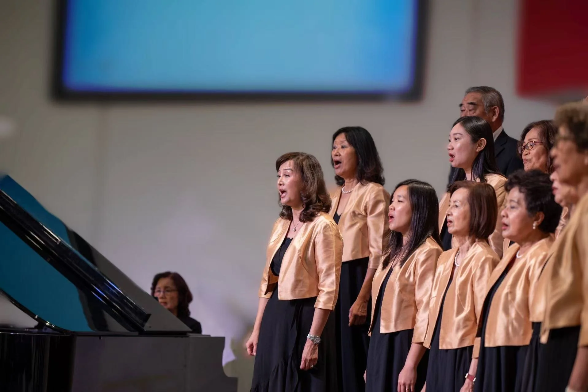 A choir of women singing in a concert hall, wearing matching gold jackets and black dresses, with a grand piano nearby.