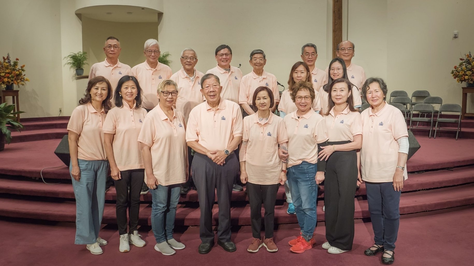 A group of 17 people, both men and women, standing together on a stage in a church setting, all wearing matching peach-colored polo shirts with a small logo. The background shows auditorium chairs and decorative floral arrangements.