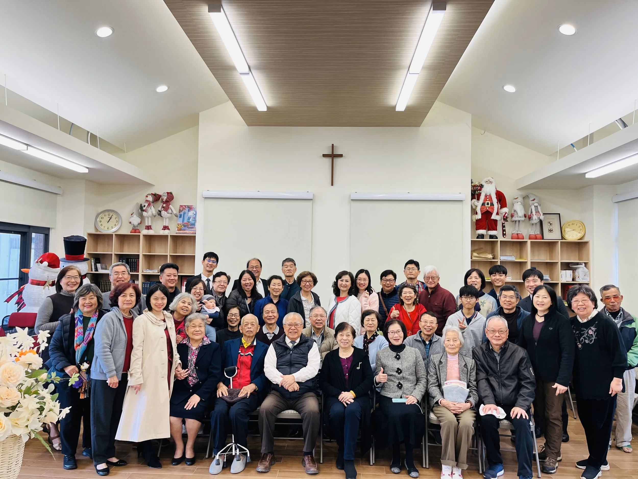 Group of people gathered in a room decorated with Christmas decorations, a cross on the wall, and Christmas figures such as Santa Claus and snowmen.