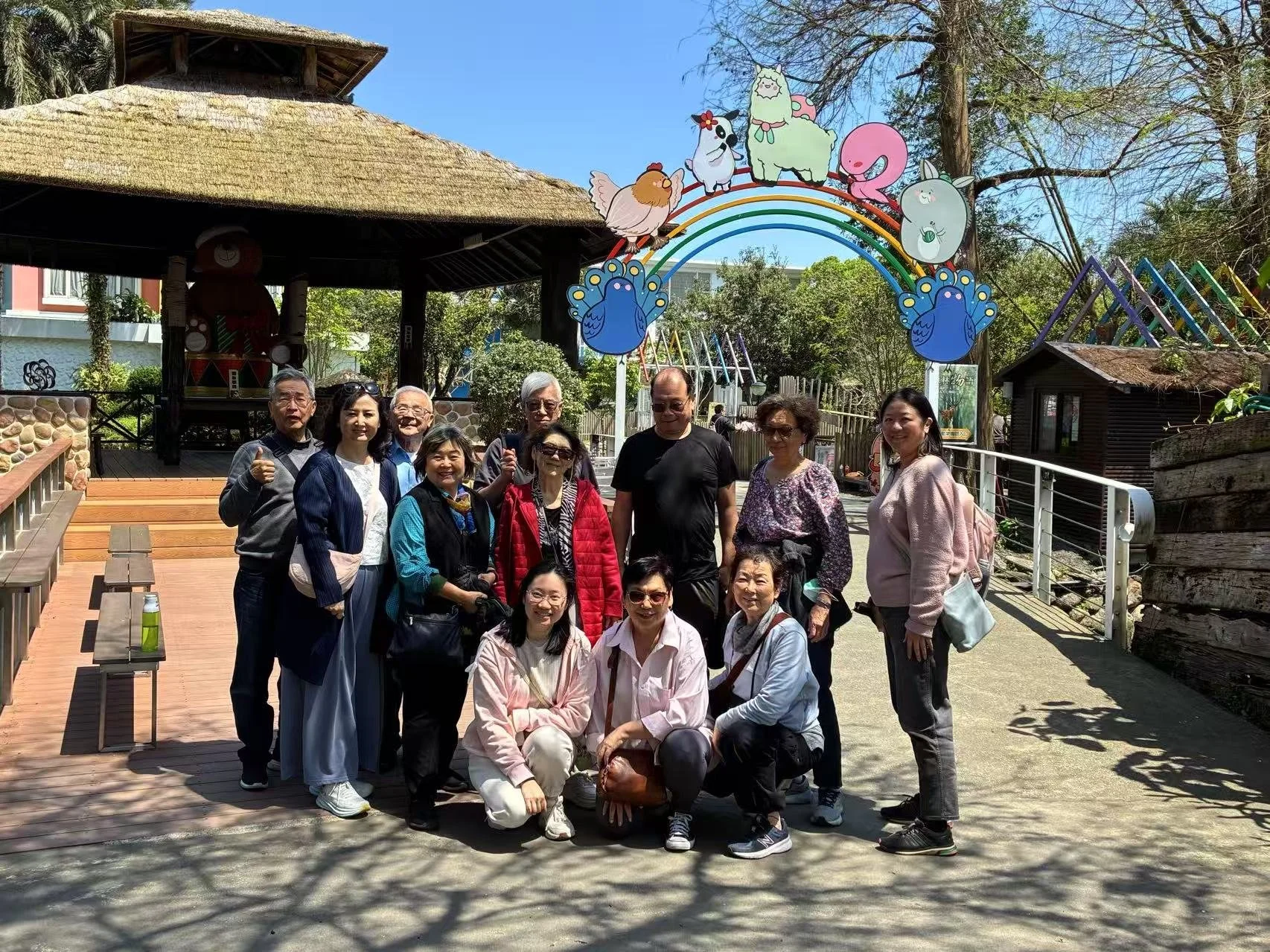 Group of 14 people smiling and posing for a photo at an amusement park with colorful animal and rainbow decorations in the background.
