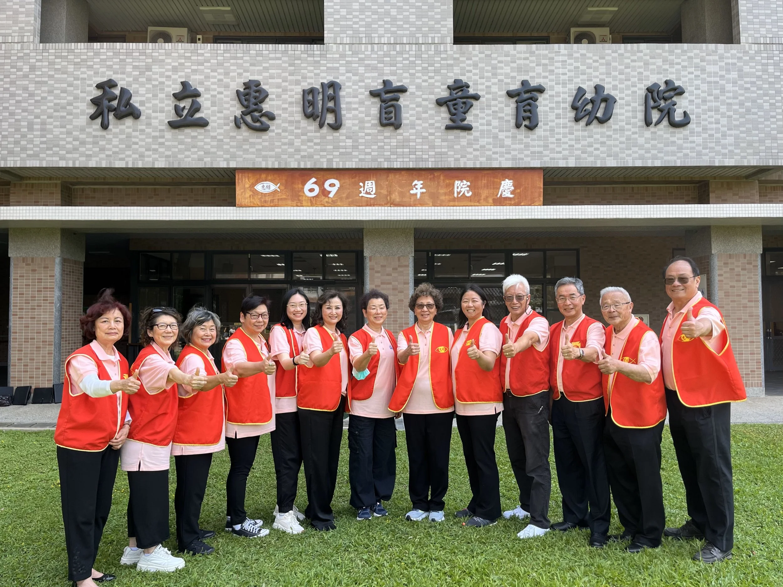 A group of fifteen elderly people standing on grass, wearing matching red vests and pink shirts, giving a thumbs-up in front of a building with Chinese characters and a sign that reads '69'. they are smiling at the camera.