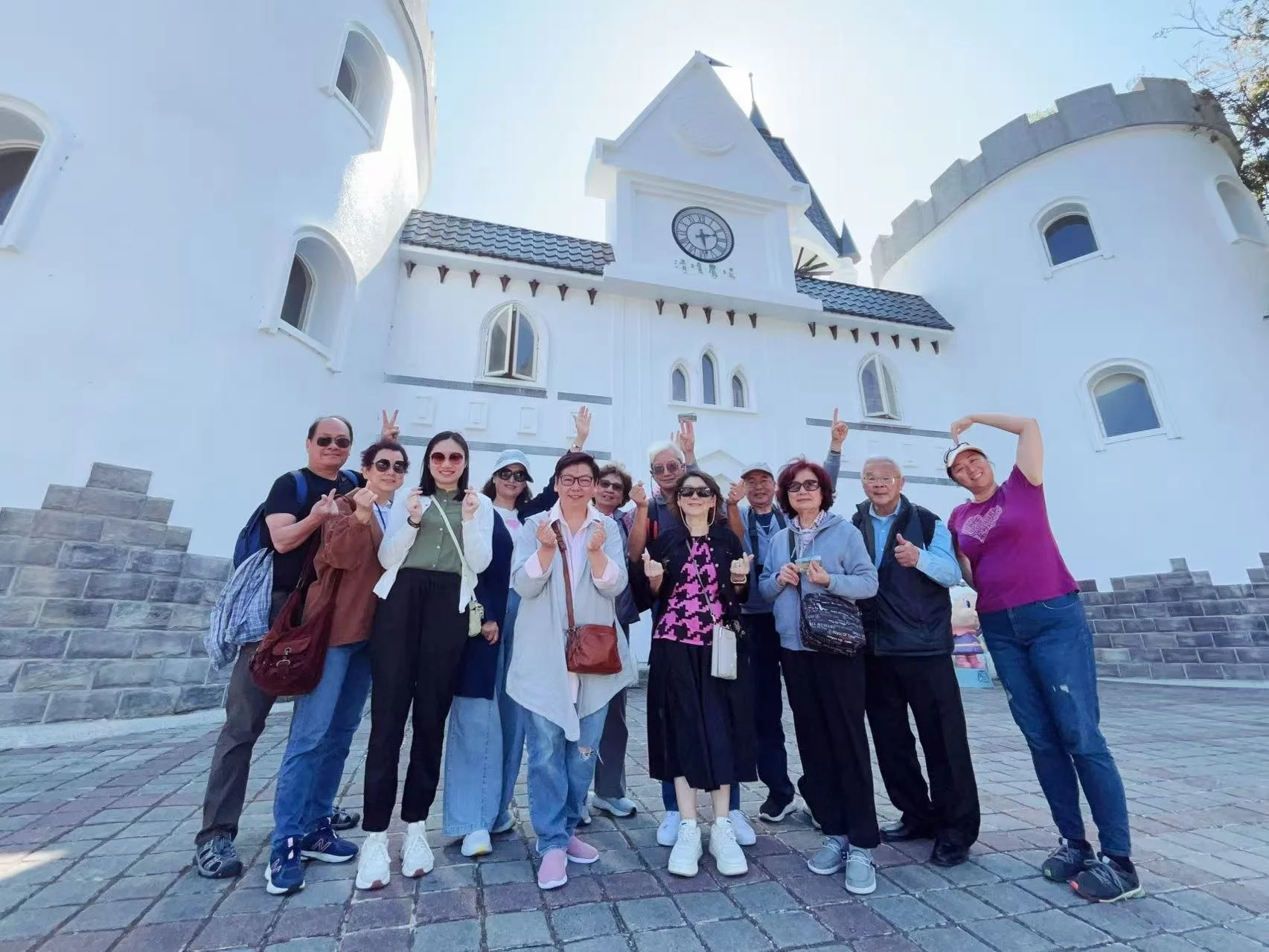 Group of people standing in front of a white castle-like building with a clock, posing for a photo and making peace signs.
