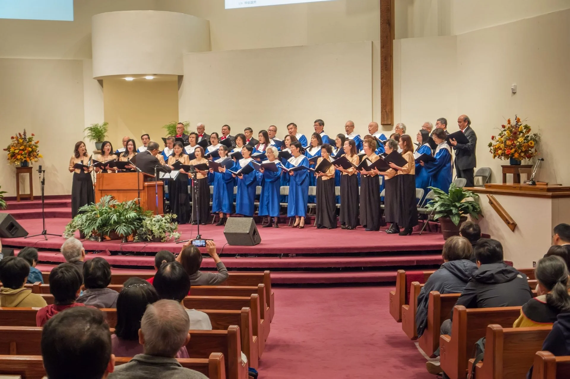 Choir performing on stage in a church or auditorium with audience seated and watching.