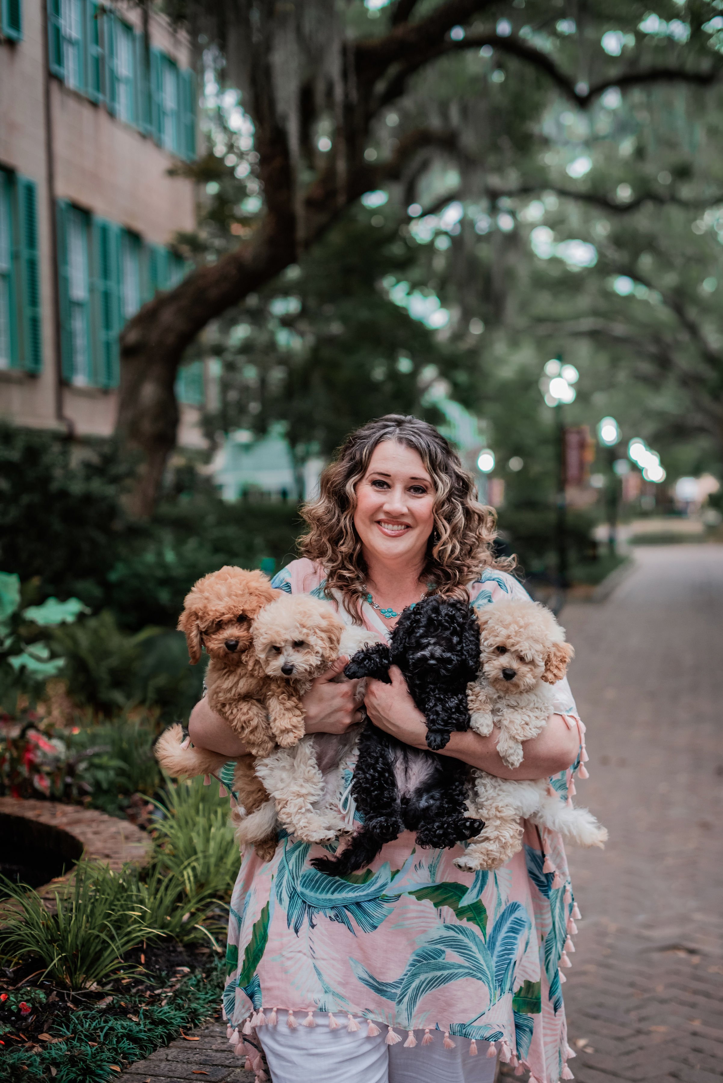 Woman holding four puppies on a brick path with trees and buildings in the background.