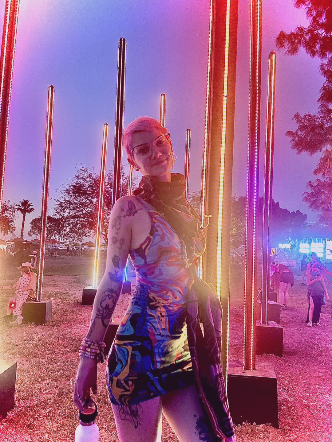 A person with short hair is in a dark room illuminated by pink and purple lights, standing against a patterned wall.