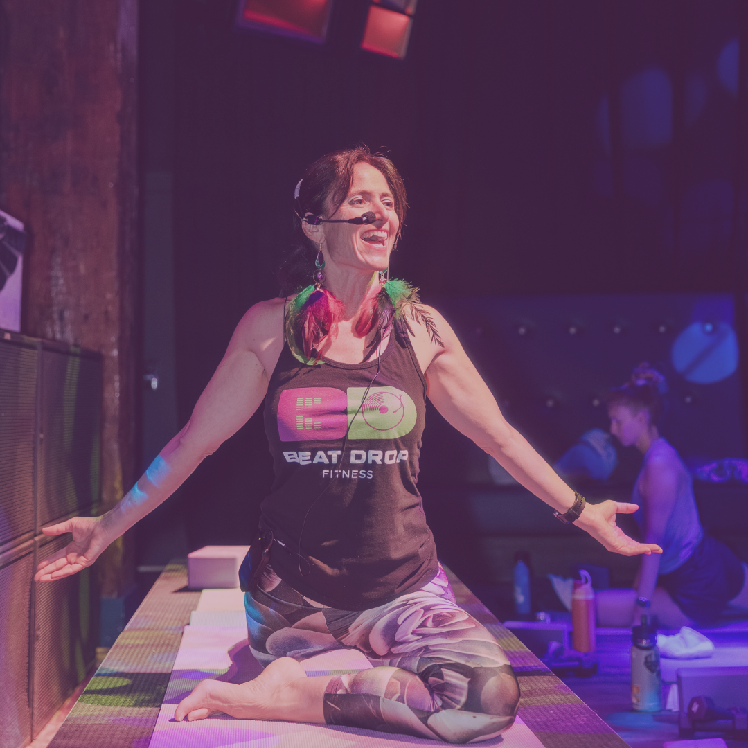 A person with short hair is in a dark room illuminated by pink and purple lights, standing against a patterned wall.