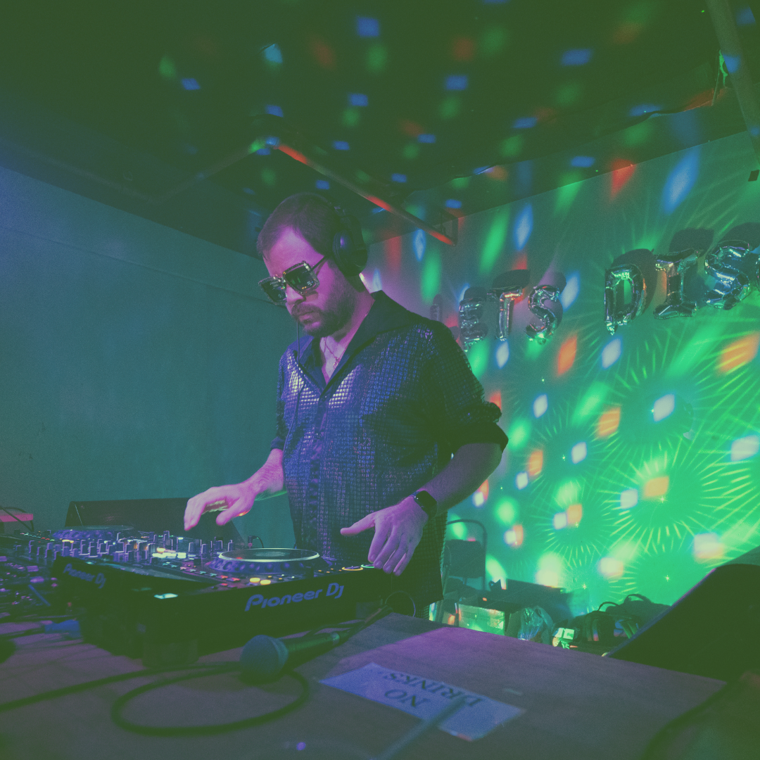 A silhouette of a person performing as a DJ on stage, illuminated by blue spotlight and surrounded by dark ambient lighting and fog.