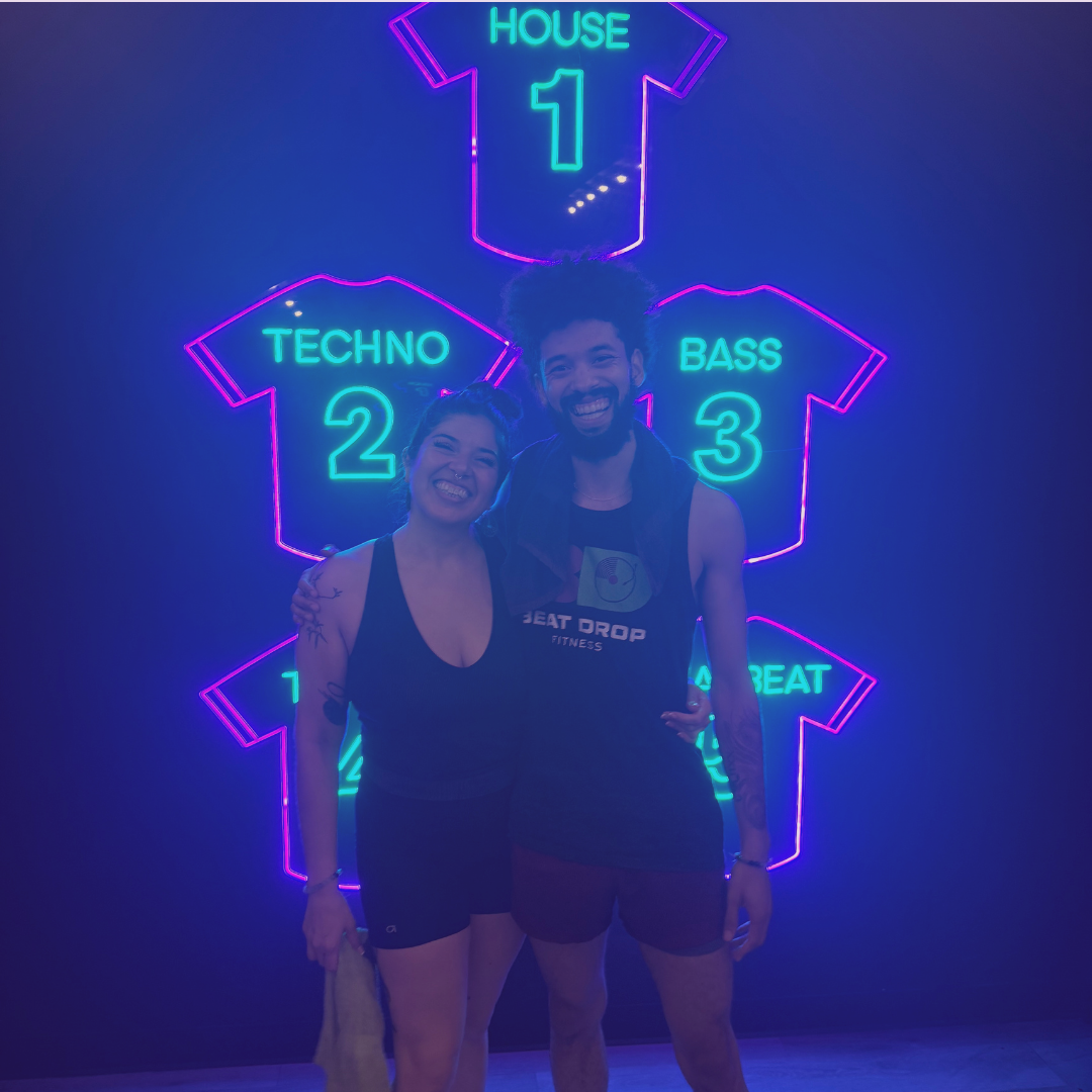 A person with short hair is in a dark room illuminated by pink and purple lights, standing against a patterned wall.