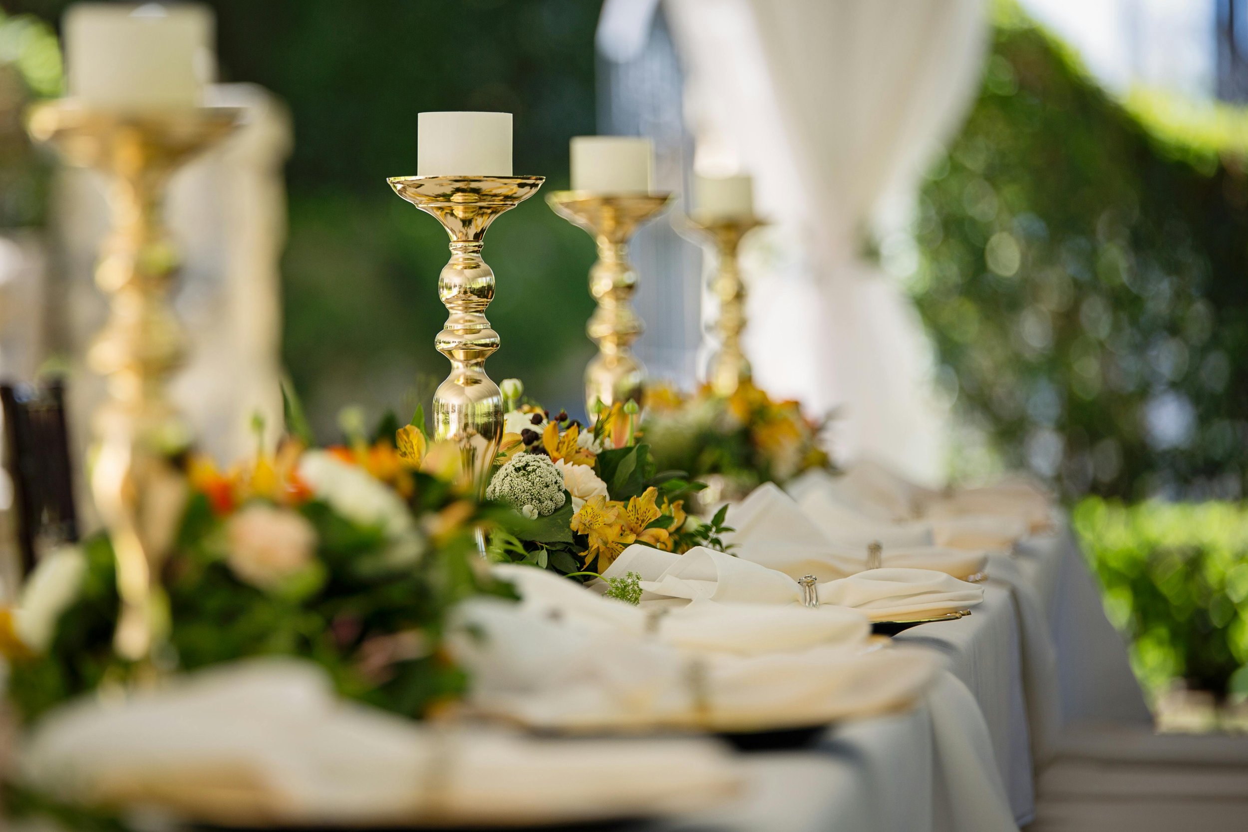 A table setting with tall gold candle holders, white candles, floral arrangements, and white napkins on a white tablecloth, outdoors with greenery in the background.