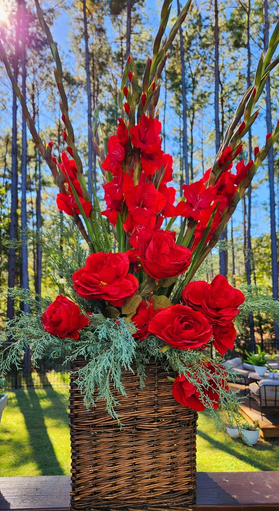 A vibrant bouquet of red flowers, including gladiolus and roses, arranged in a wicker basket outside on a wooden surface with tall trees and a clear blue sky in the background.