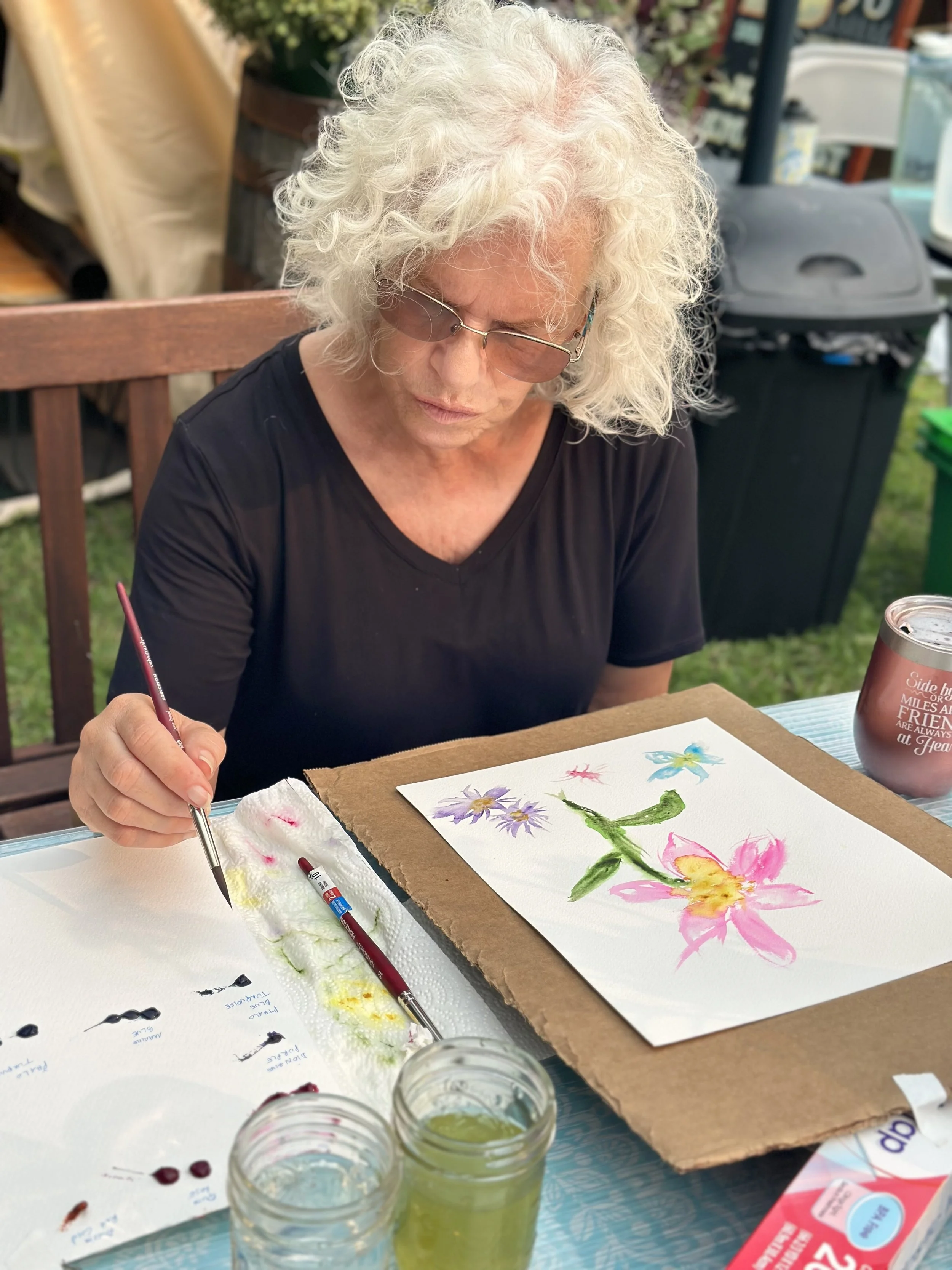 An elderly woman with curly white hair and glasses painting a colorful floral watercolor on paper outdoors. She is seated at a table with art supplies, including jars of water, paintbrushes, and a palette, with a decorated background.