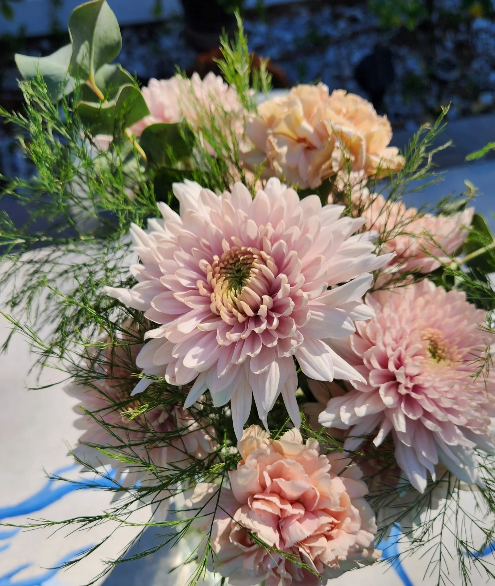 A bouquet of light pink chrysanthemums and carnations with greenery in natural sunlight.