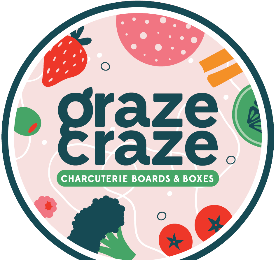 A round charcuterie board featuring assorted vegetables, strawberries, and other food items with the text "graze graze" and a green banner reading "Charcuterie Boards & Boxes."