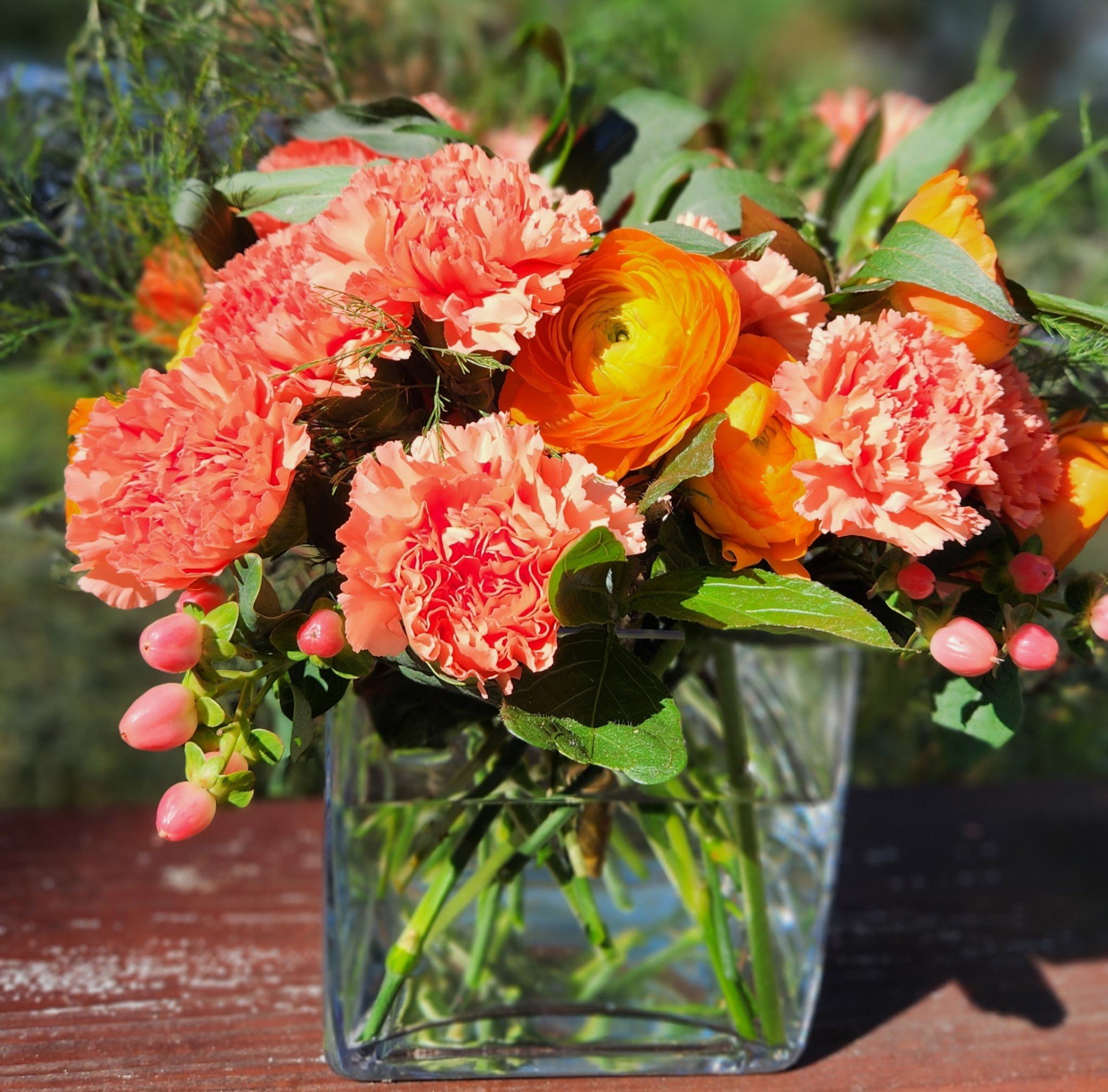 A bouquet of pink carnations and orange ranunculus flowers in a clear glass vase on a wooden surface with a blurred outdoor background.