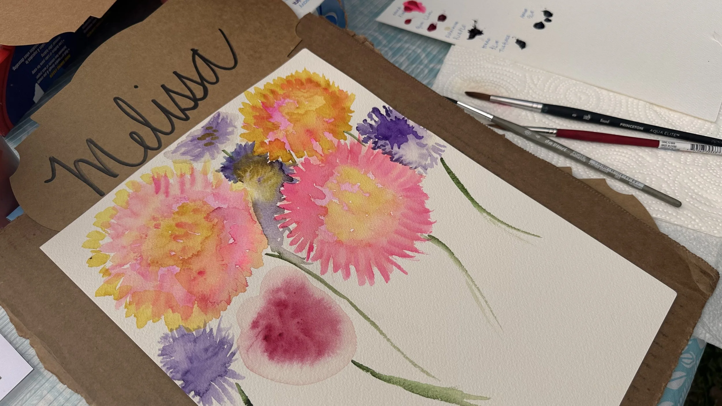 A watercolor painting of colorful flowers with pink, yellow, purple, and red blooms on textured paper, surrounded by brushes and art supplies.
