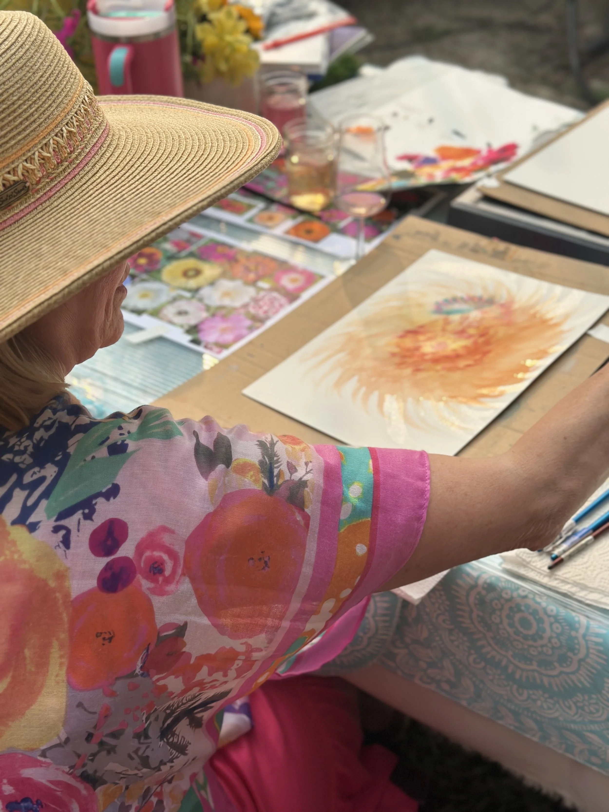 A woman at an outdoor art table, wearing a wide-brimmed straw hat and colorful floral blouse, works on a watercolor painting of a sunflower. The table is cluttered with paintings, sketches, brushes, glasses of water, and art supplies, with additional