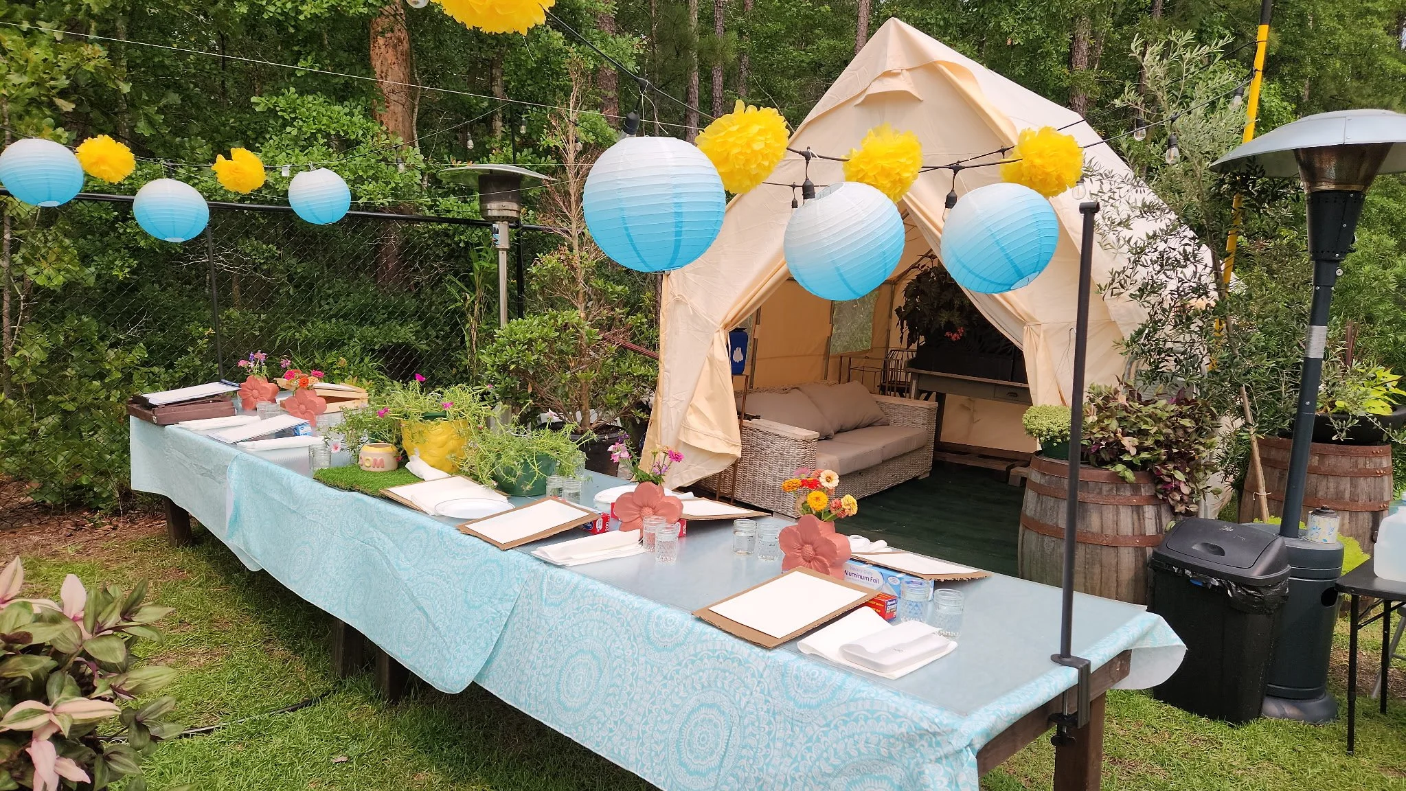 Outdoor celebration setup with a long table covered in a light blue tablecloth, decorated with pink and yellow flowers in green pots, surrounded by green plants and trees. Above, hanging blue paper lanterns and yellow pom-pom decorations. In the back