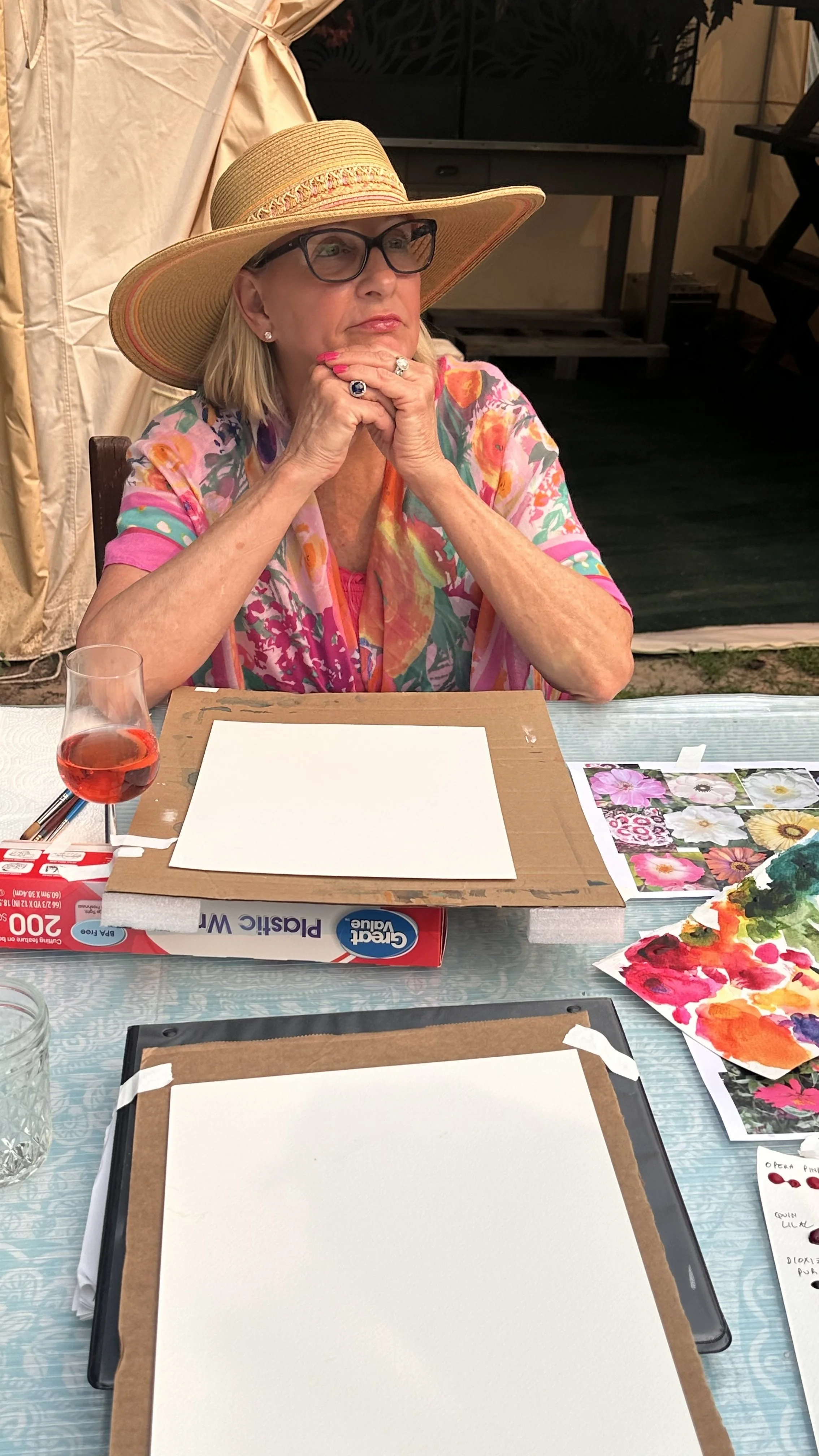 An elderly woman wearing a wide-brimmed straw hat, glasses, and a colorful floral top, sitting at a table with art supplies, papers, and a glass of red drink, in an indoor setting.