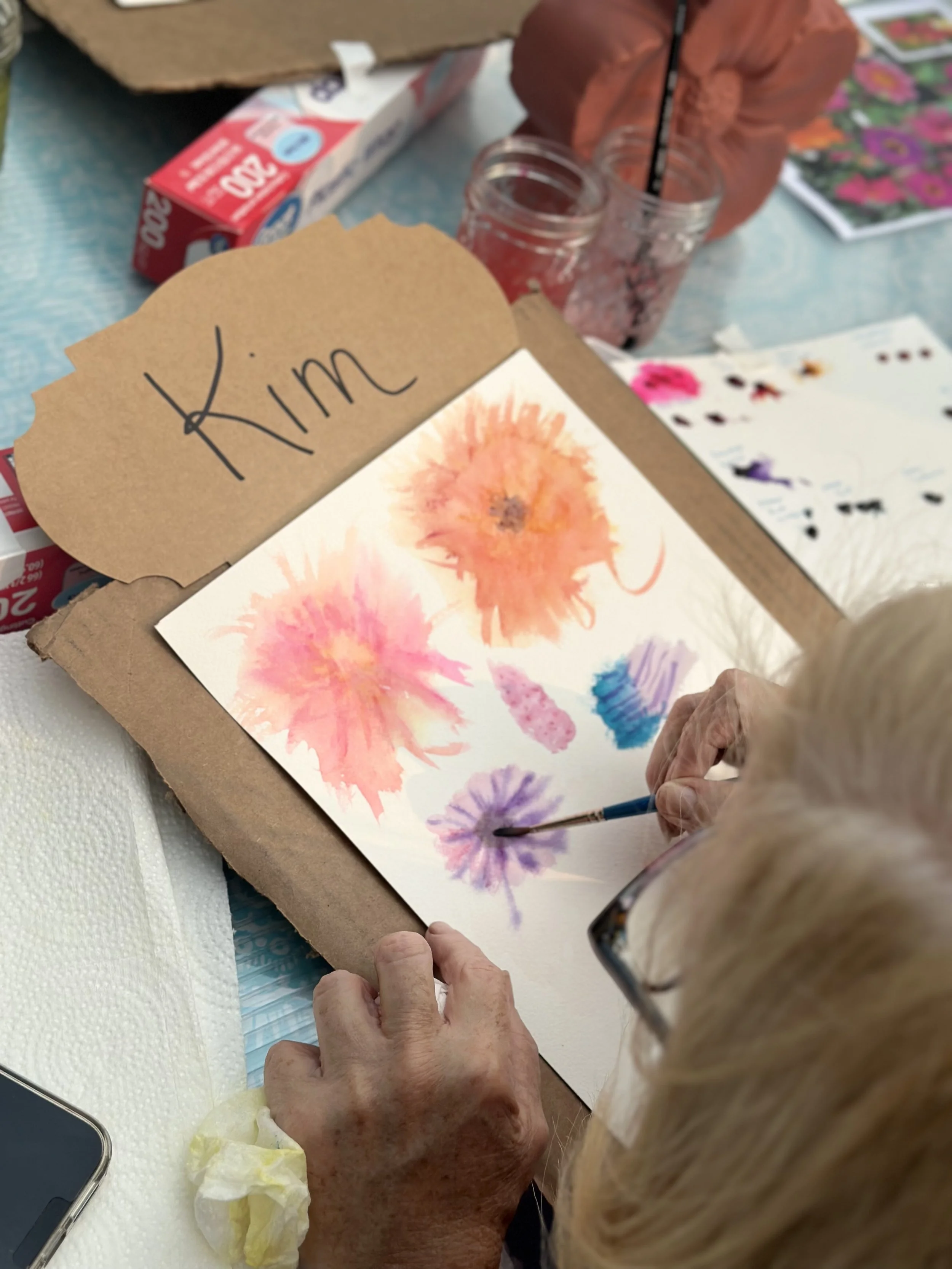 Close-up view of an elderly woman painting watercolor flowers on paper at an art table, with a brown paper sign labeled 'Kim' in the background and various art supplies nearby.