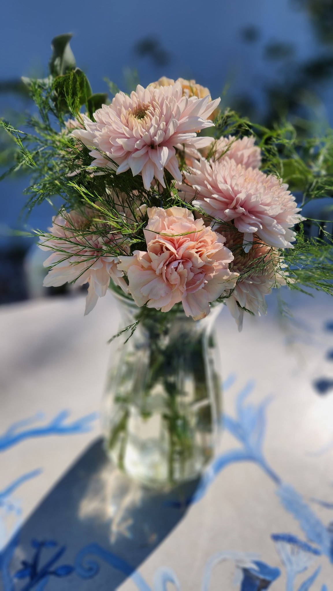 A glass vase with pink and white flowers and green foliage sitting on a table with a patterned shadow on the tablecloth.