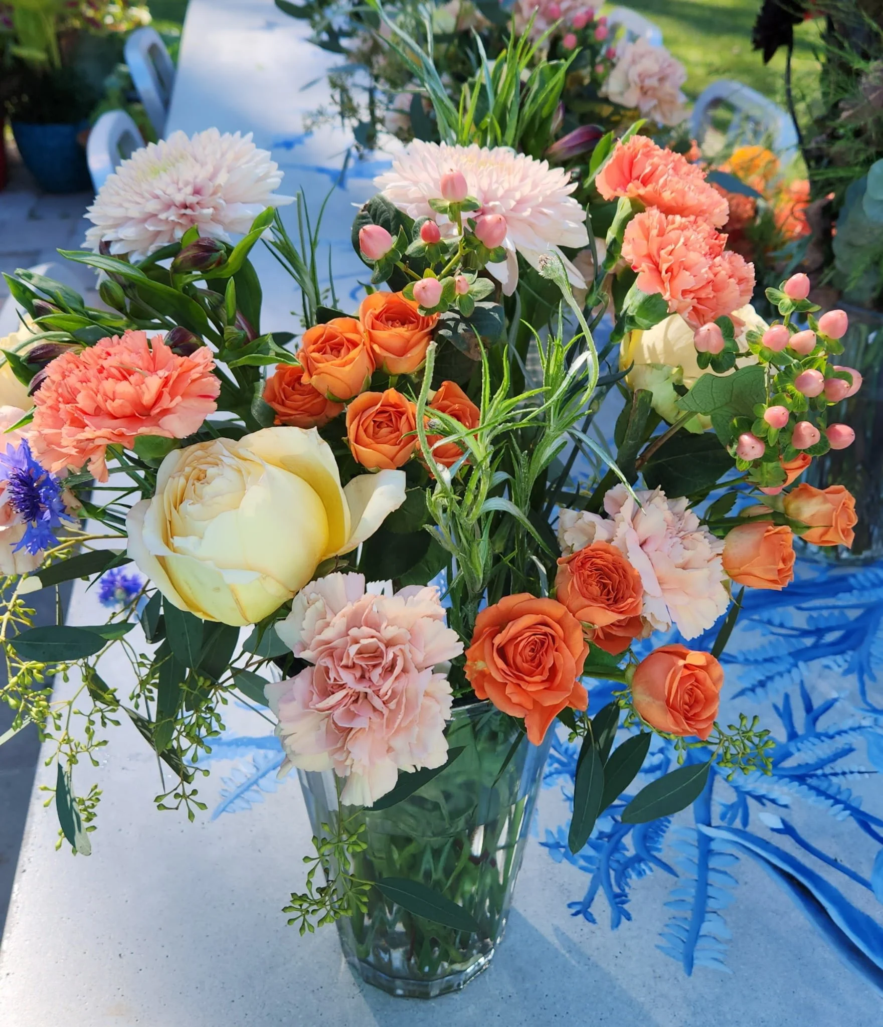 A glass vase filled with a colorful flower arrangement, including cream-colored peonies, peach and orange roses, pink carnations, blue cornflowers, and assorted greenery. The vase is on a white table outdoors with a blurred background.